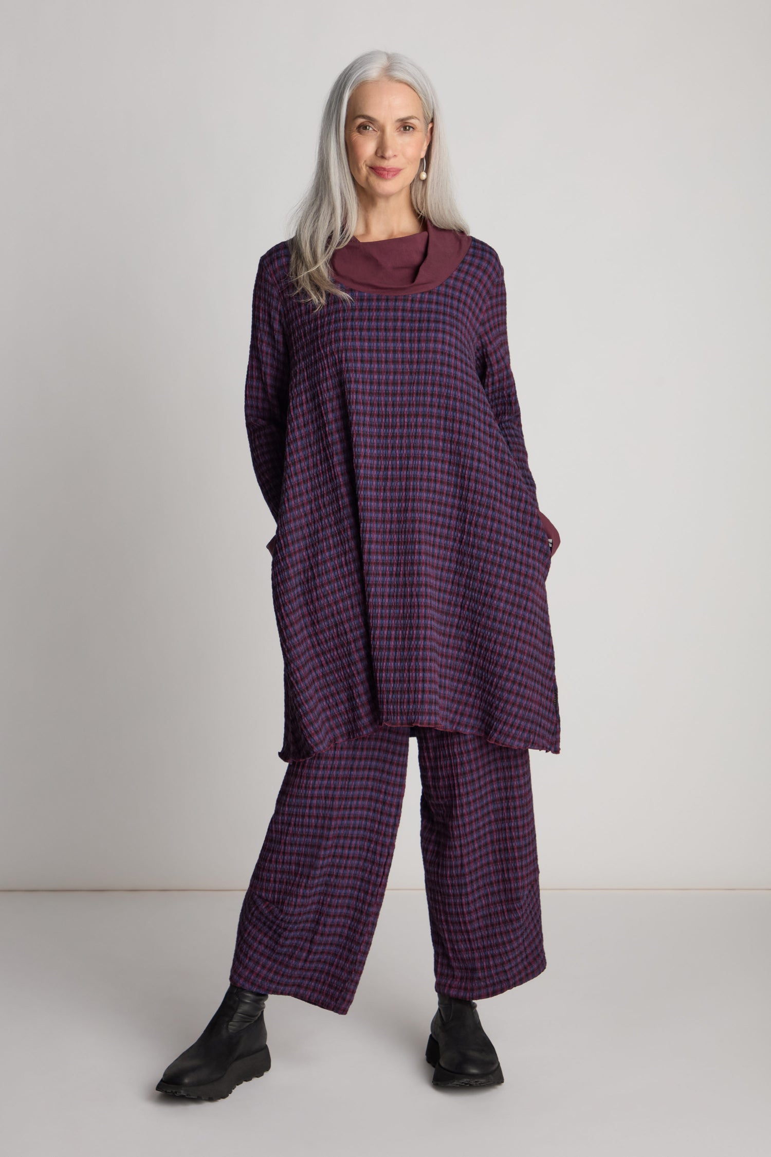 A woman with long gray hair is wearing a Luukaa Satin Cowl Check Tunic and matching pants, paired with black shoes, standing against a plain white background. This ensemble exemplifies sustainable fashion at its finest.