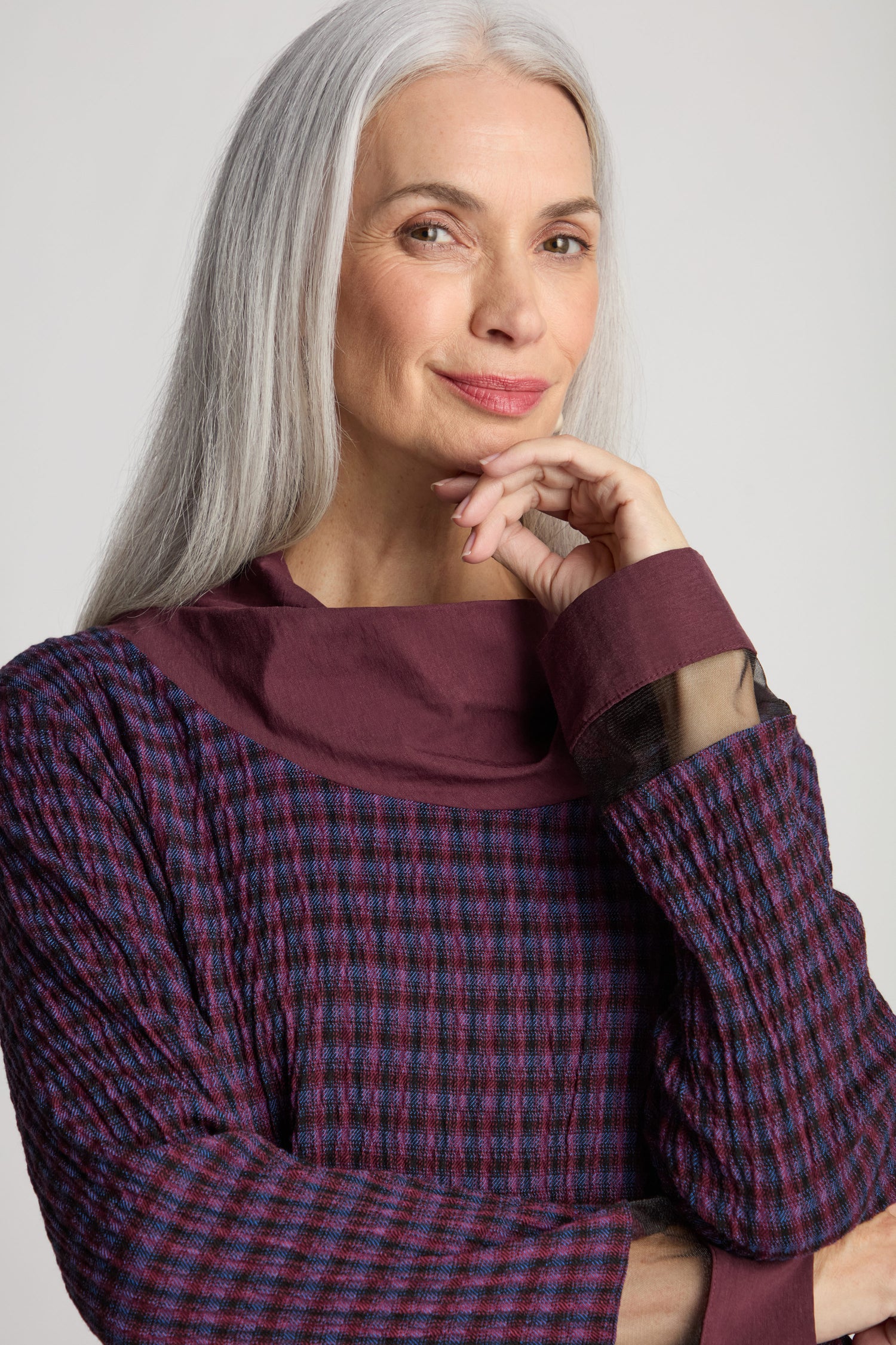 A woman with long gray hair and a thoughtful expression, wearing Luukaa's Satin Cowl Check Tunic, looks at the camera.