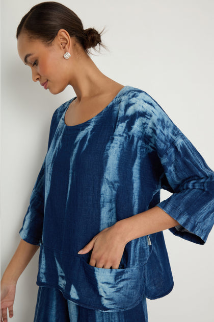 A woman models the Soft Denim Two-Pocket Top, a blue tie-dye cotton denim long-sleeve shirt with a boxy silhouette, standing sideways against a plain light background.