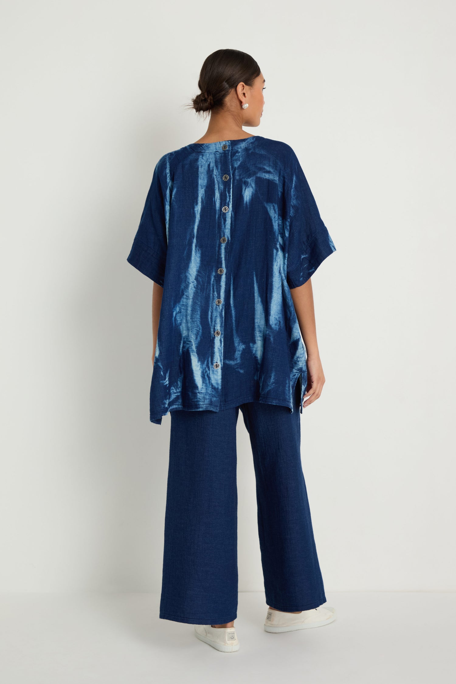 A person stands facing away, wearing the Soft Denim Button-Back Tunic with short sleeves and a longline silhouette, paired with matching wide-leg blue pants and white sneakers.