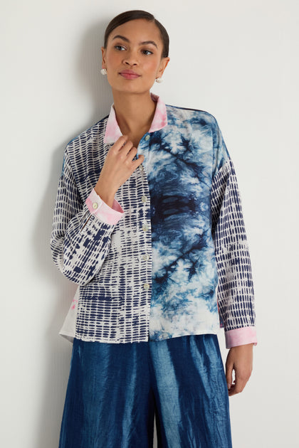 A woman stands against a white wall wearing the Floral Back Easy Shirt in blue and white with pink accents, paired with loose blue pants.