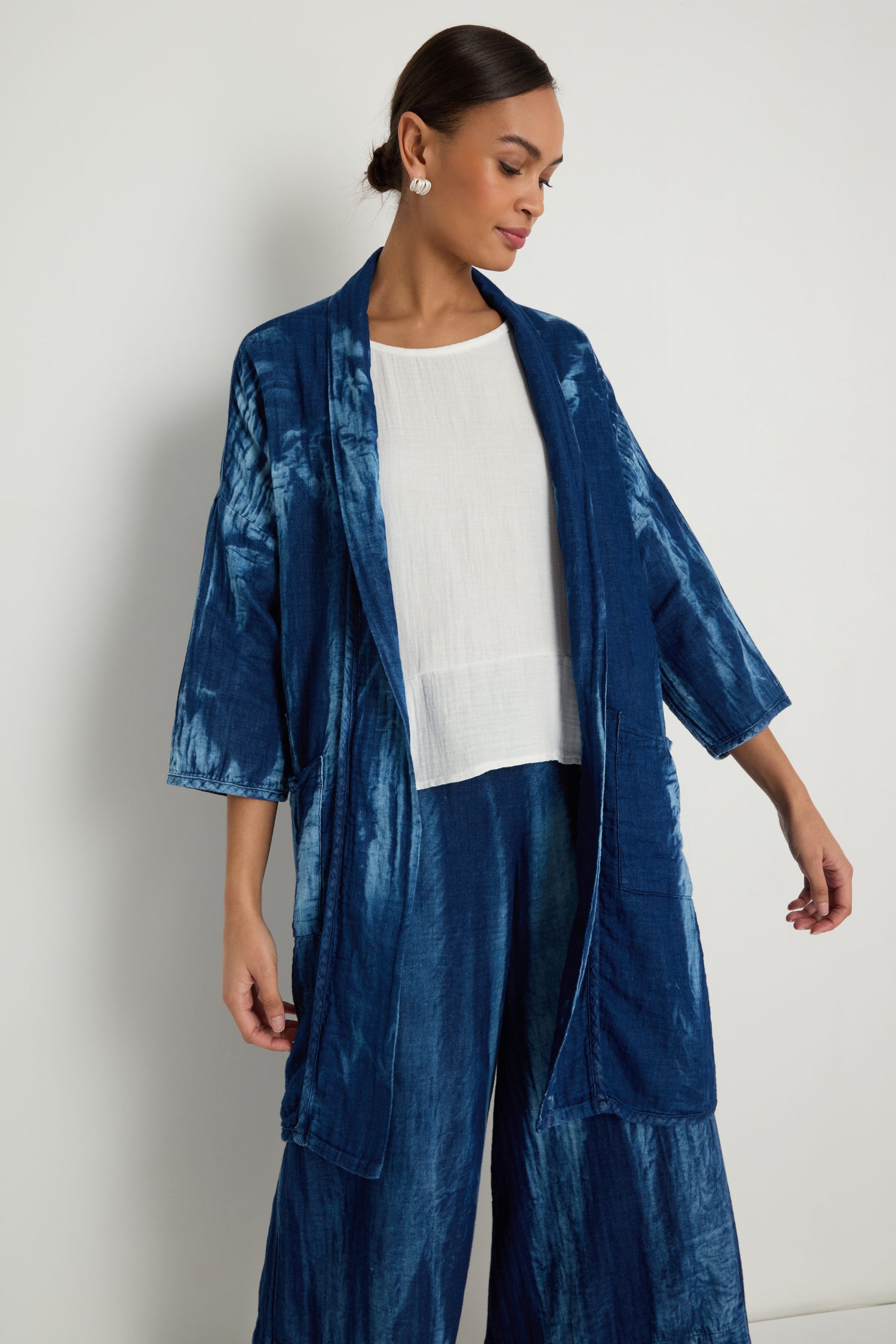 A woman stands against a plain white wall, wearing the Soft Denim Easy Jacket in a relaxed blue tie-dye style over a white top, paired with matching pants.