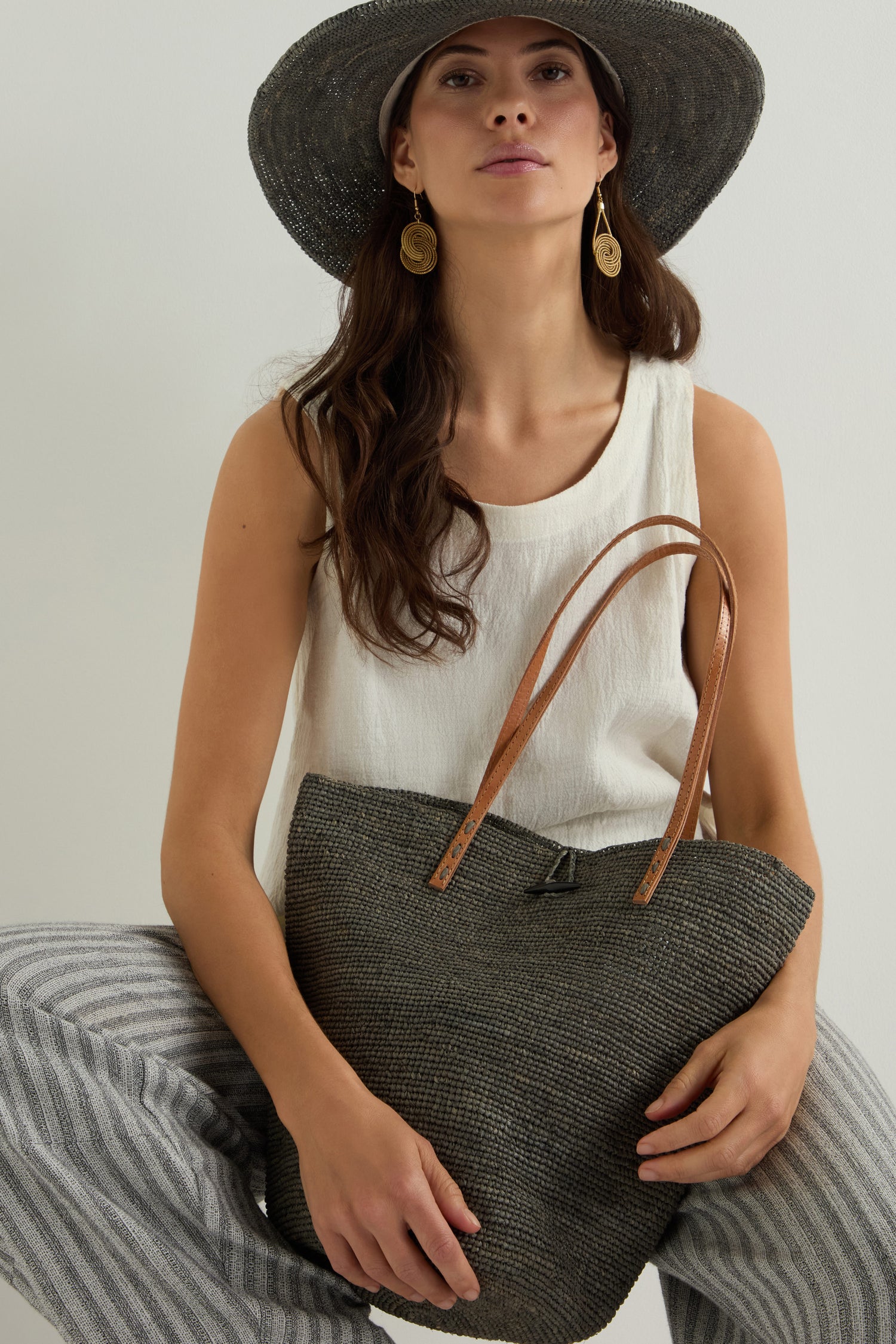 A woman in a white top, striped pants, and large sun hat displays the Ilana Raffia Basket, highlighting its handwoven raffia design and roomy interior with leather straps.