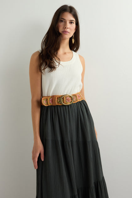 A woman in a sleeveless white top and a long dark skirt, accented by the Cinzoa Raffia Belt and an earring, stands against a plain light wall.