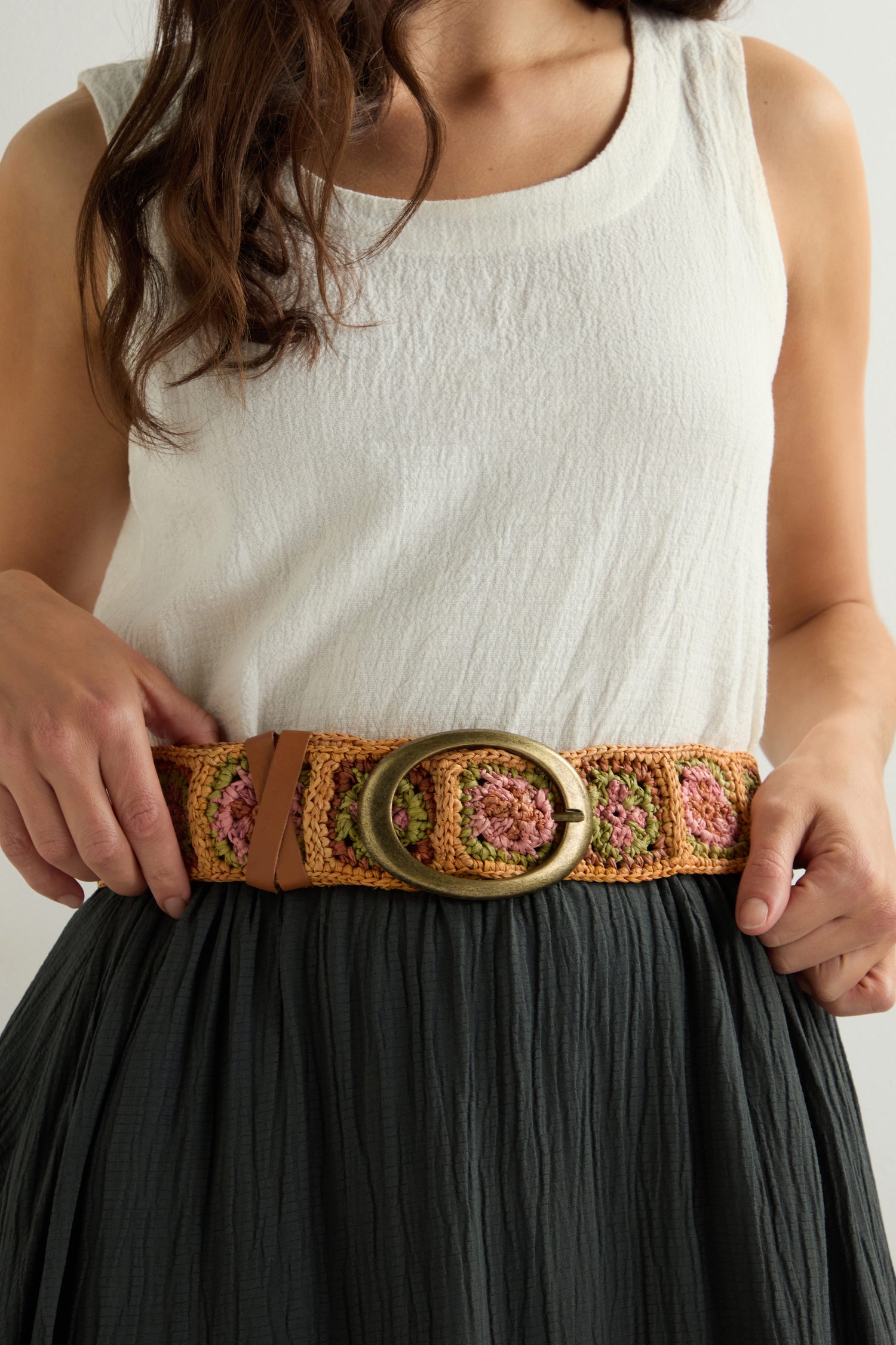 A person in a white sleeveless top and dark skirt adjusts their Cinzoa Raffia Belt with a round brass buckle.