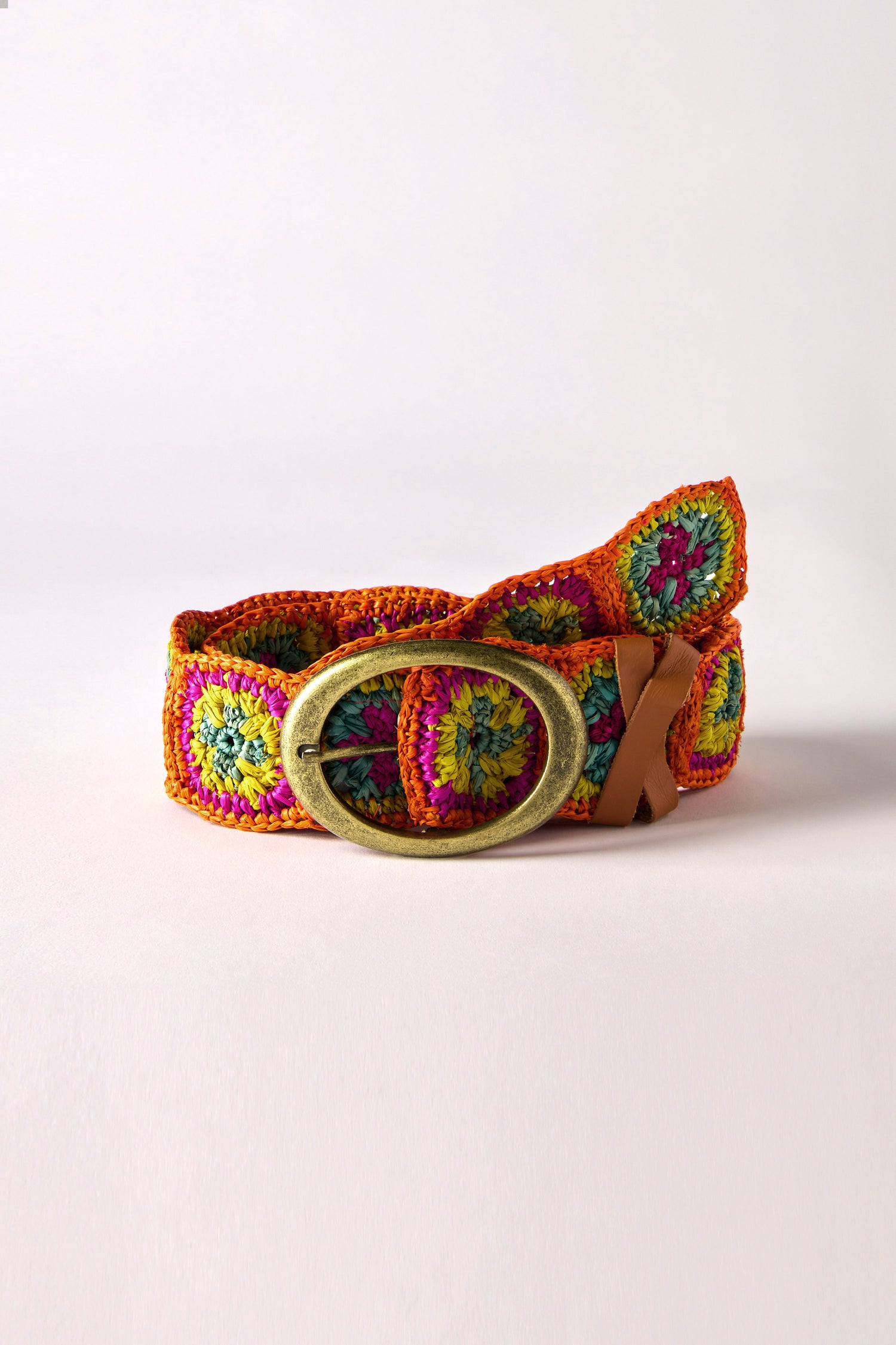 The Rainbow Cinzoa Raffia Belt is a boho-chic accessory featuring vibrant circular patterns in green, yellow, and pink crocheted design, complete with a large oval brass buckle.