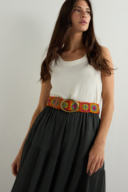 A woman in a white sleeveless top and dark skirt stands against a plain light background, accentuated by a boho-chic touch: the Rainbow Cinzoa Raffia Belt with vibrant circular patterns, adding versatility and flair to her ensemble.