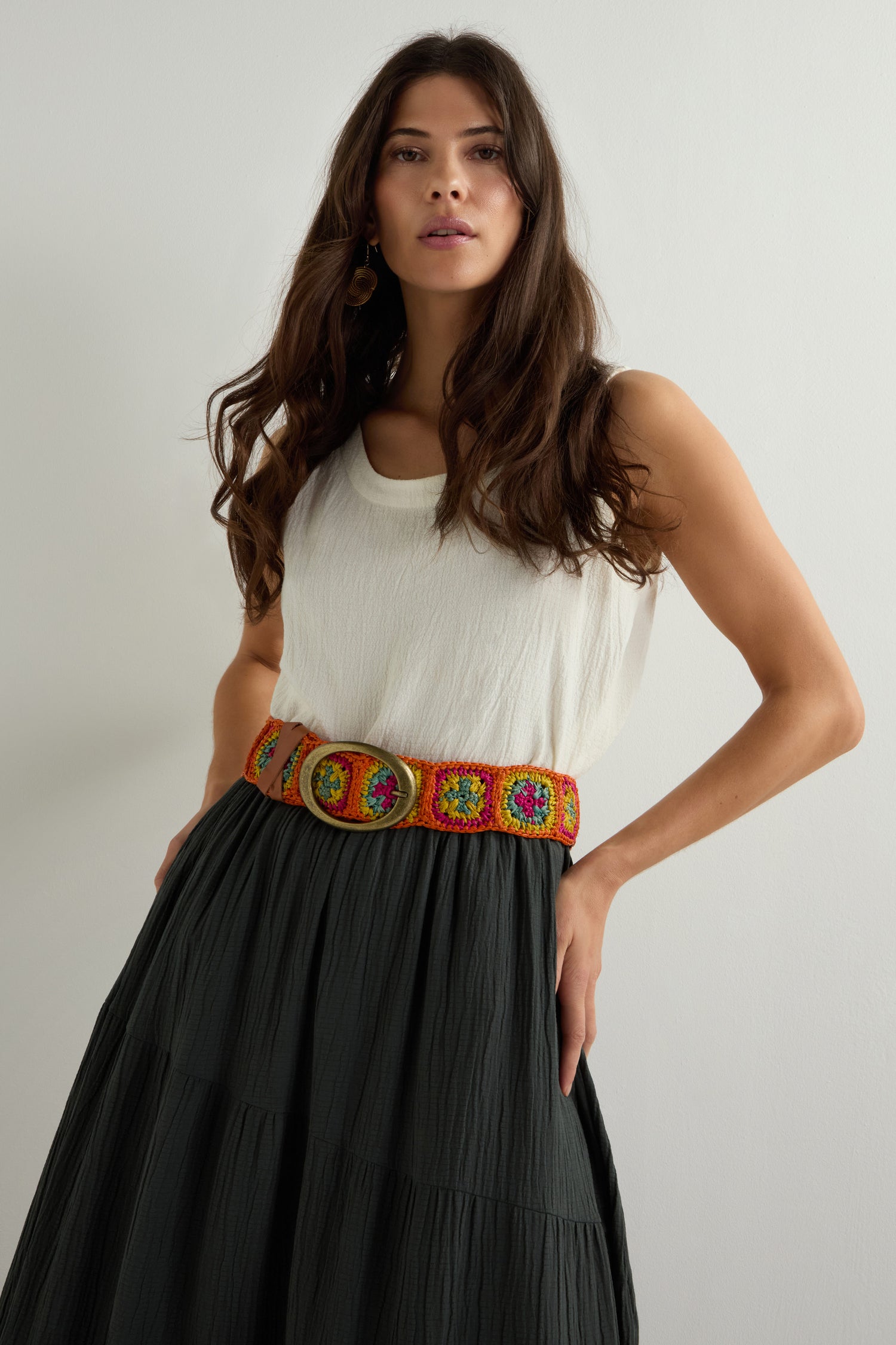 A woman with long hair wears a white tank top and black skirt, accessorized with a Rainbow Cinzoa Raffia Belt, adding boho-chic flair as she stands against a plain backdrop.