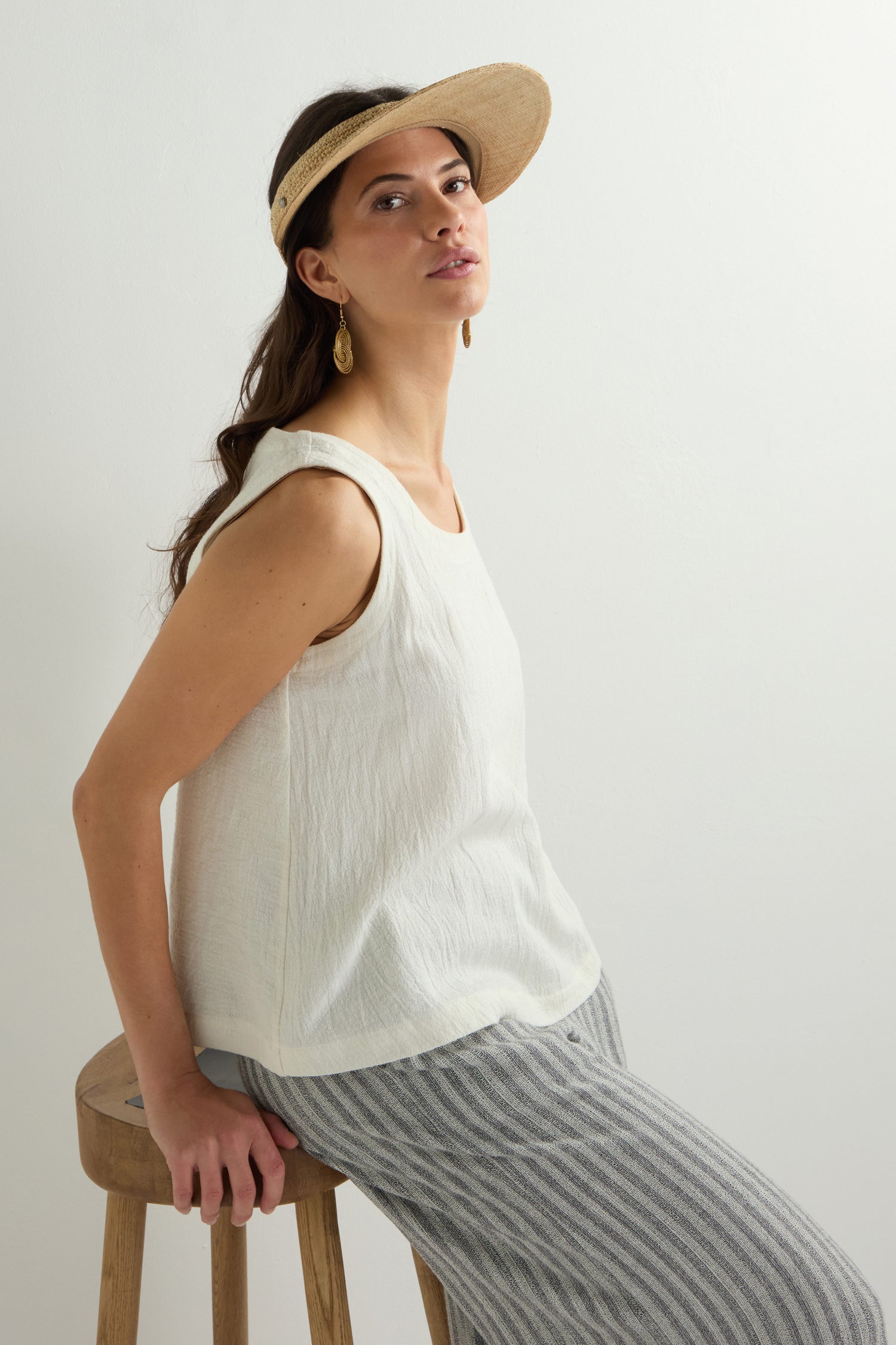 A person sits on a wooden stool, wearing a white sleeveless top and striped pants. They don the Garossoa Visor by Le Voyage en Panier for sun protection, along with drop earrings, facing slightly to the side against a plain background.