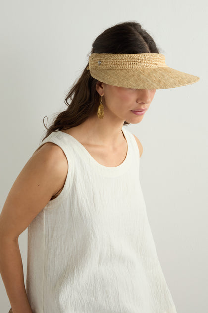 A person with long brown hair wears a tan Garossoa Visor and white sleeveless top, accessorized with gold earrings, embodying style and sun protection. The simple light background emphasizes their understated elegance.