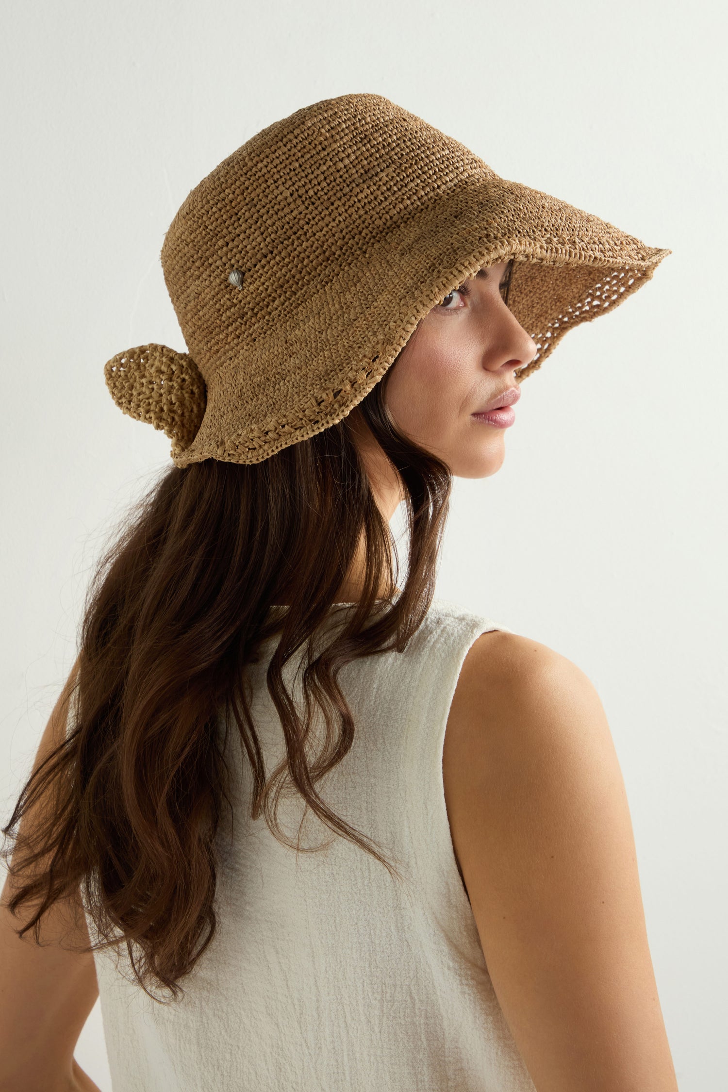 A woman embodies effortless summer style in a sleeveless white top and the Chapeau Noloa, a natural woven straw sun hat, as she gazes over her shoulder against a light backdrop.