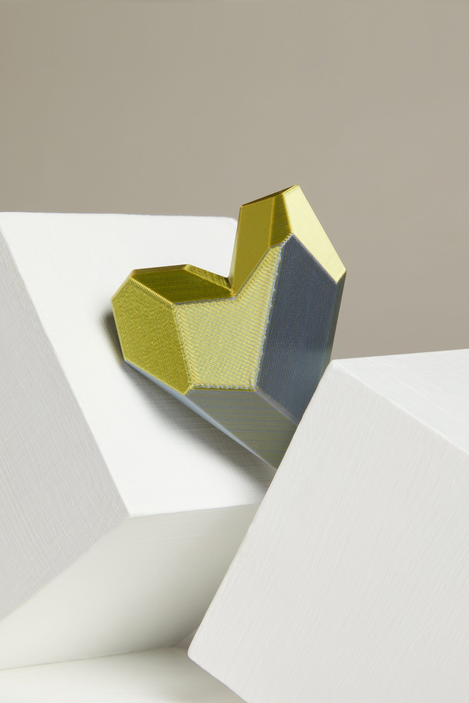 The 3D Printed Heart Brooch showcases a geometric heart design, made using advanced 3D printing techniques, with gold and gray facets set between two white blocks on a beige background.