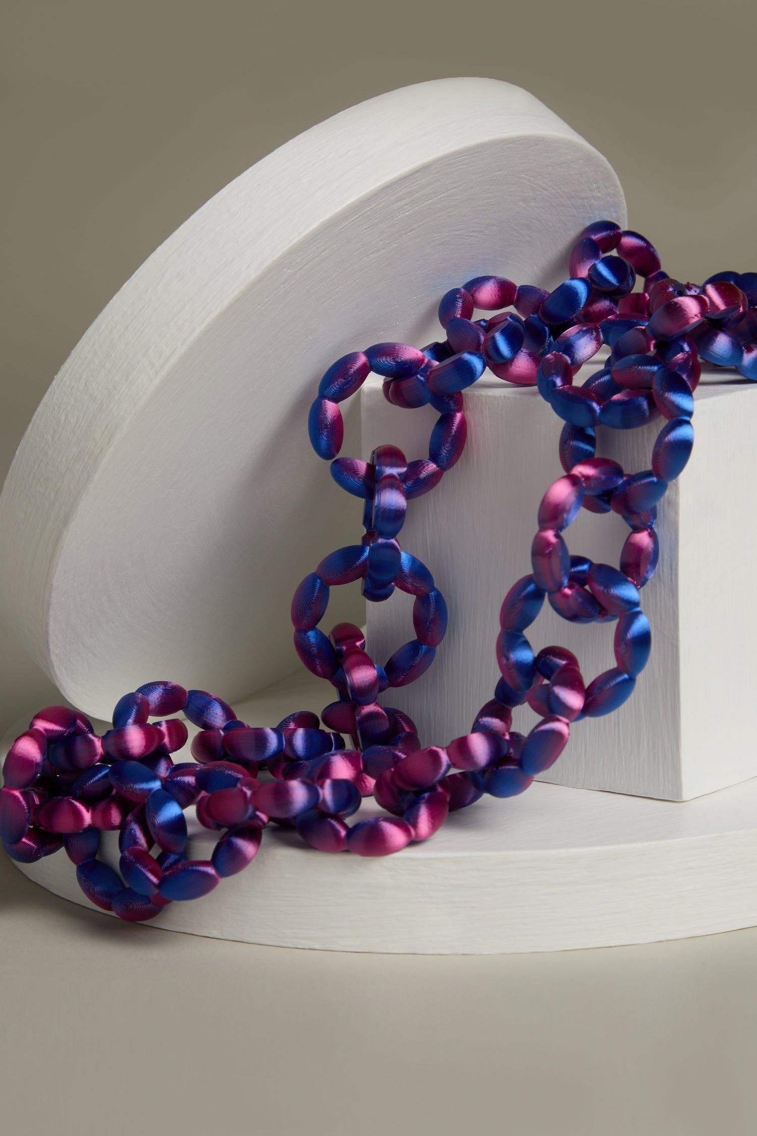 The ultra-lightweight 3D Printed Links Necklace by Maison 203, with its interlocking oval links in shades of purple and blue, is elegantly displayed draped over a white circular stand against a neutral background.