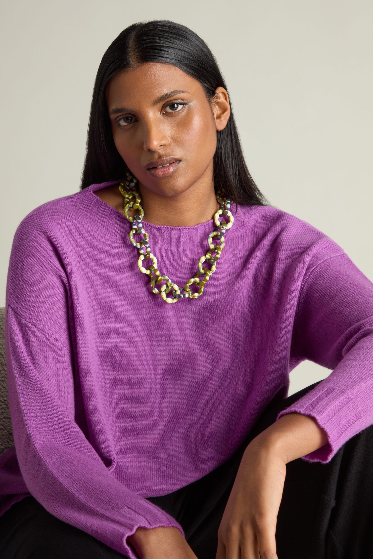 A woman in a purple sweater wearing the 3D Printed Links Necklace from Maison 203, known for its ultra-lightweight design, sits gracefully against a neutral background. The chunky chain necklace adds an airy elegance to her look.