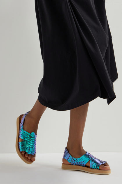 A person elegantly walks on a light surface, wearing a long black skirt and Frida Woven Leather Sandals. These colorful, handcrafted shoes from Mexico feature a flat sole and bring artisanal charm.
