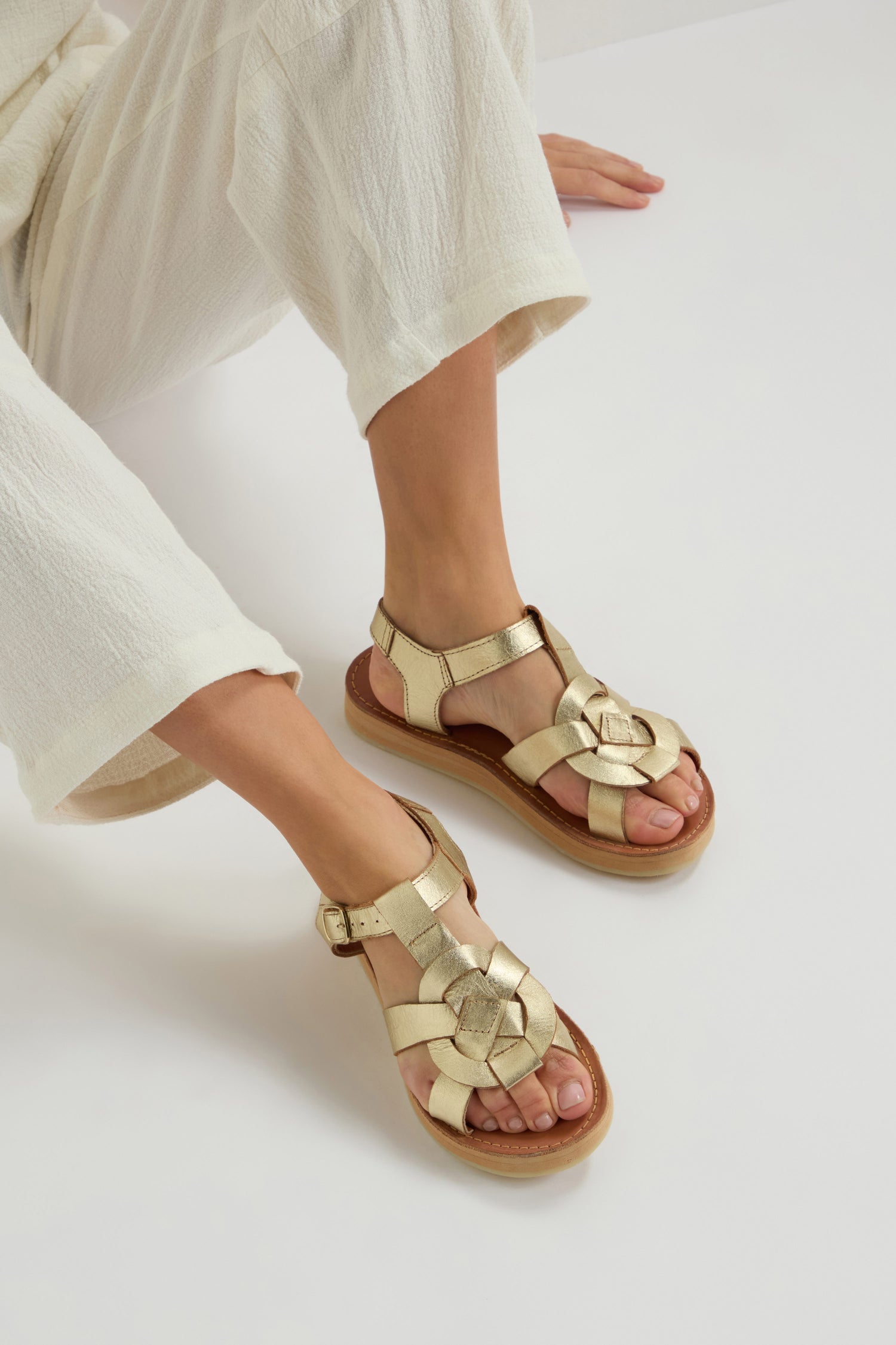 A person sits on a white surface, wearing white pants with Tulum Leather Buckle Sandals featuring gold open-toe design, woven straps, and a cushioned sole for comfort.