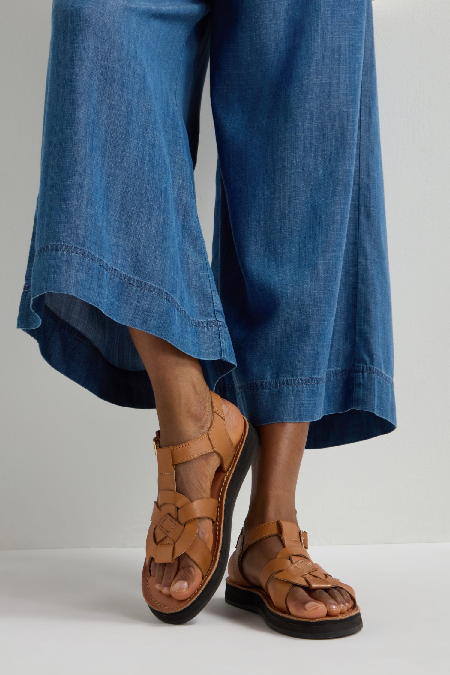 A person in wide-leg denim and Tulum Leather Buckle Sandals stands against a white background, showcasing style with cushioned sole comfort.