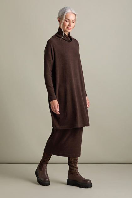 A person with grey hair wearing a luxurious fleece knit Allai Rib Neck Soft Jersey Dress, a brown skirt, and brown boots is standing against a plain light background.