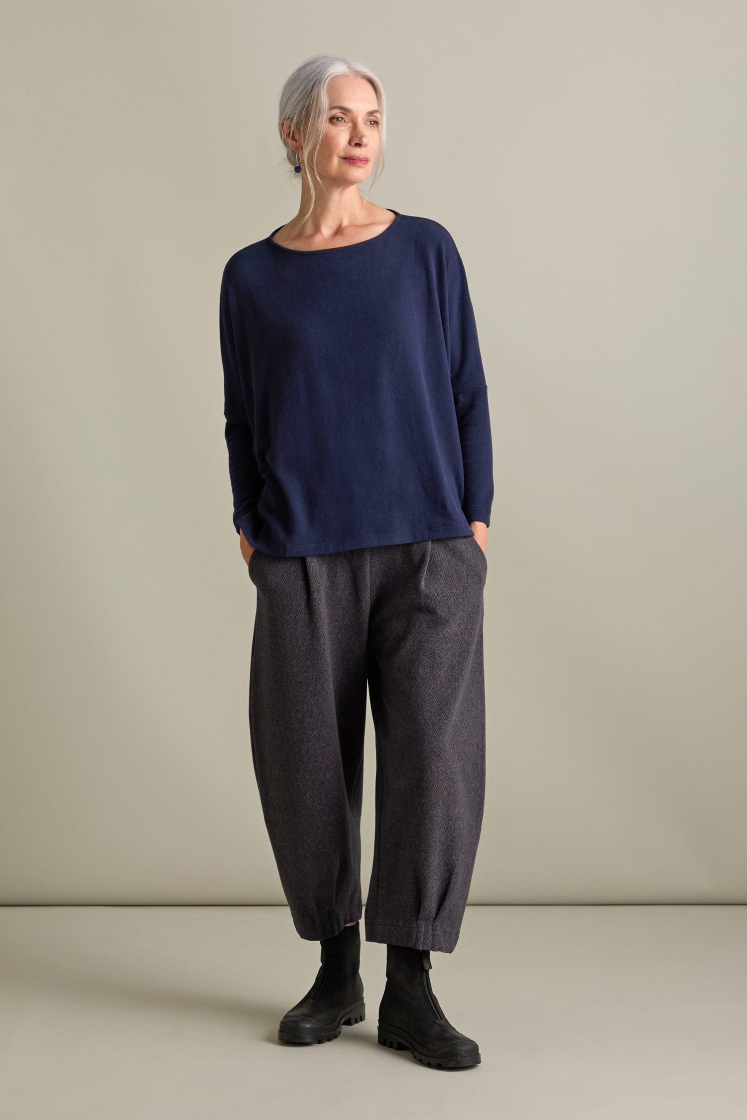 A woman with gray hair is standing in a relaxed pose, wearing a navy long-sleeve top and dark Biano Jersey Jacquard trousers that showcase Italian craftsmanship. She has her hands in her pockets, black boots on her feet, and is smiling slightly.