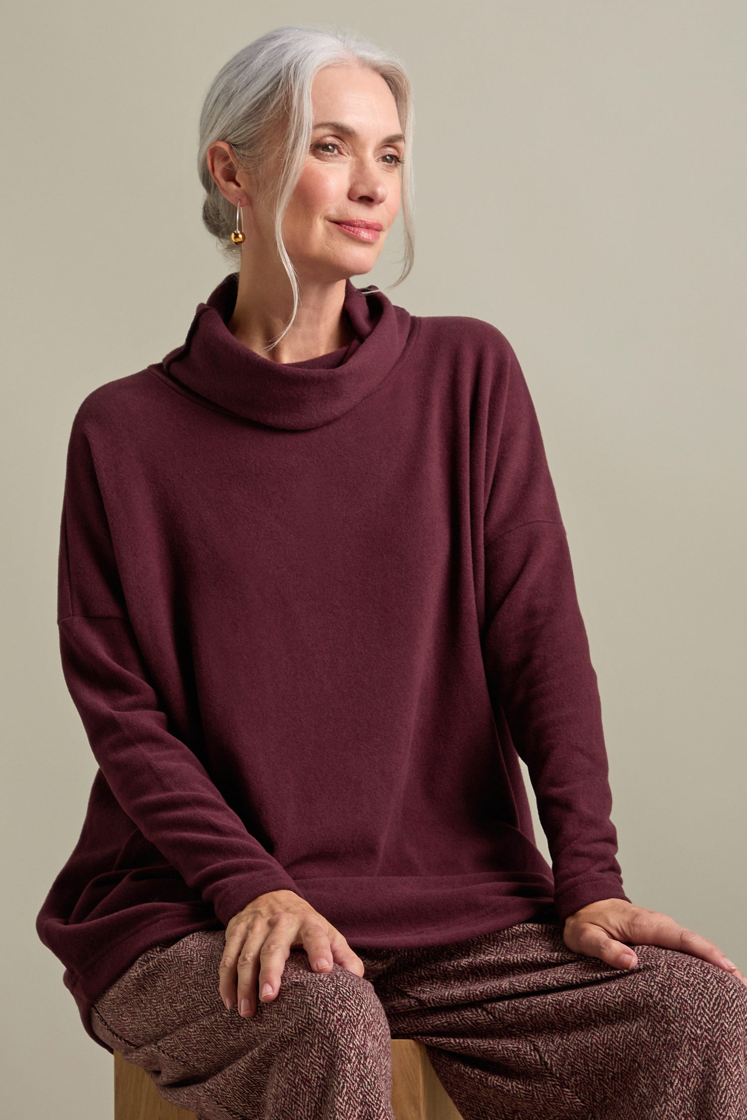 A woman with gray hair is sitting on a stool, wearing an oversized maroon Bill Roll Neck Soft Jersey Top and patterned pants.