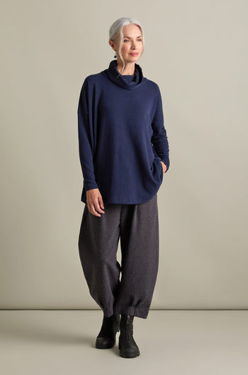 An older woman with grey hair stands relaxed against a neutral backdrop. She wears a Bill Roll Neck Soft Jersey Top in navy blue, loose grey pants, and black boots. She has one hand in her pocket and is looking off to the side.