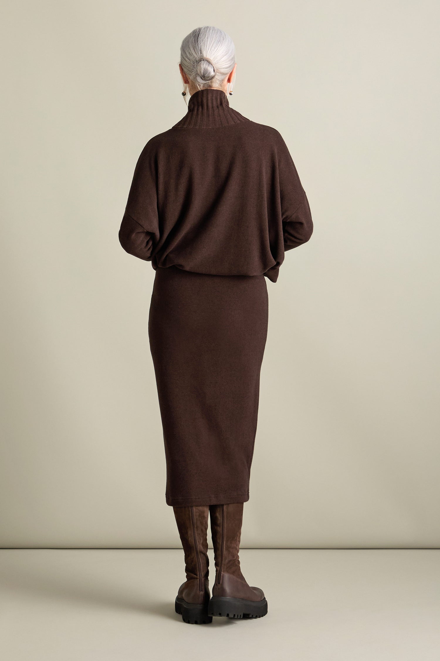 A person with gray hair styled in a bun is seen from the back, wearing a brown turtleneck sweater, matching Lilla Soft Jersey Tube Skirt, and knee-high brown boots. They stand against a plain, light-colored background, embodying the elegant simplicity of Autumn/Winter '24 fashion.