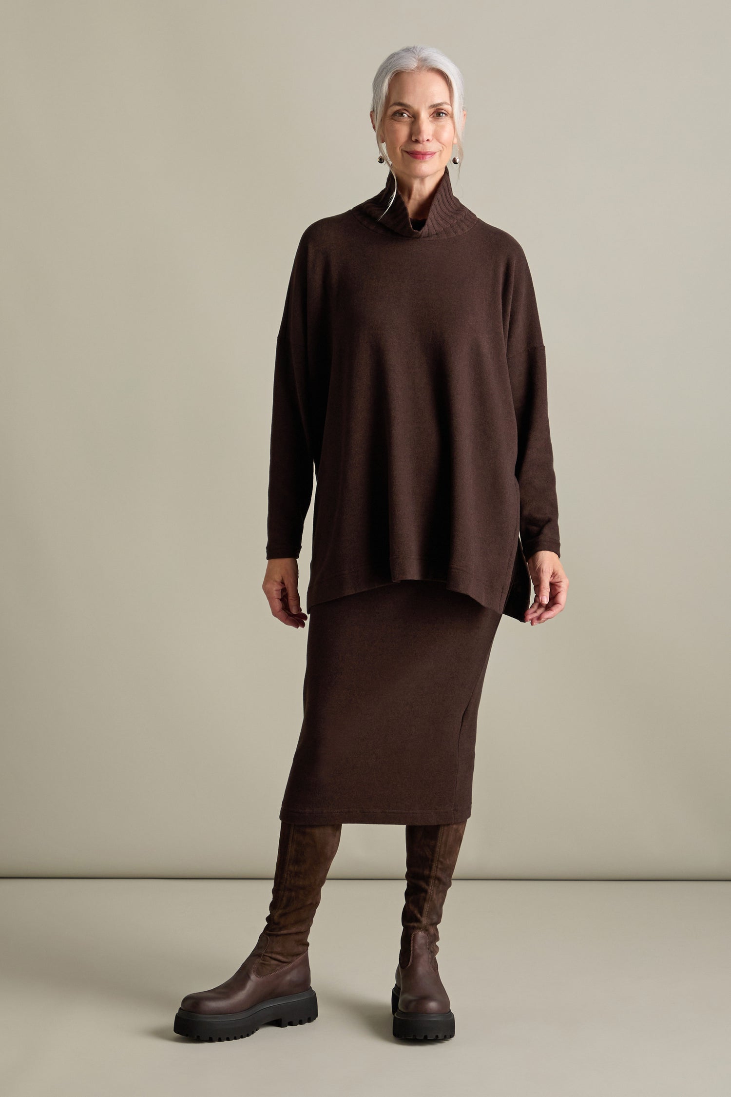 A woman with gray hair stands against a plain background wearing a long brown sweater, a matching Lilla Soft Jersey Tube Skirt from Mama b, and brown knee-high boots, embodying the cozy style of Autumn/Winter '24.