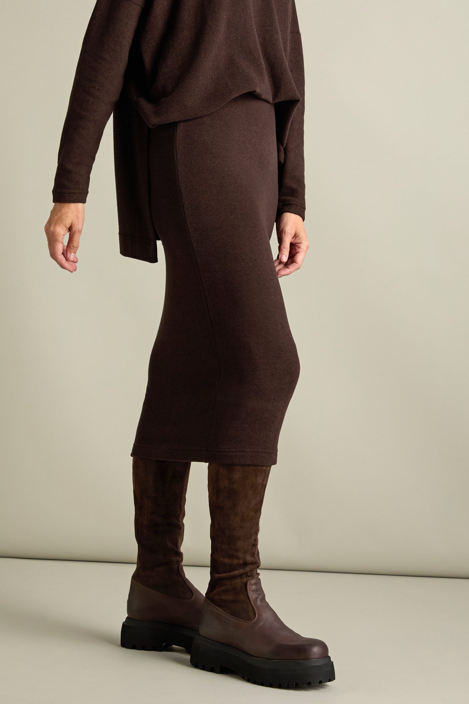 Person wearing a brown long-sleeve top and the Lilla Soft Jersey Tube Skirt in brown paired with dark brown knee-high boots, standing against a plain backdrop. Perfect for Autumn/Winter '24.