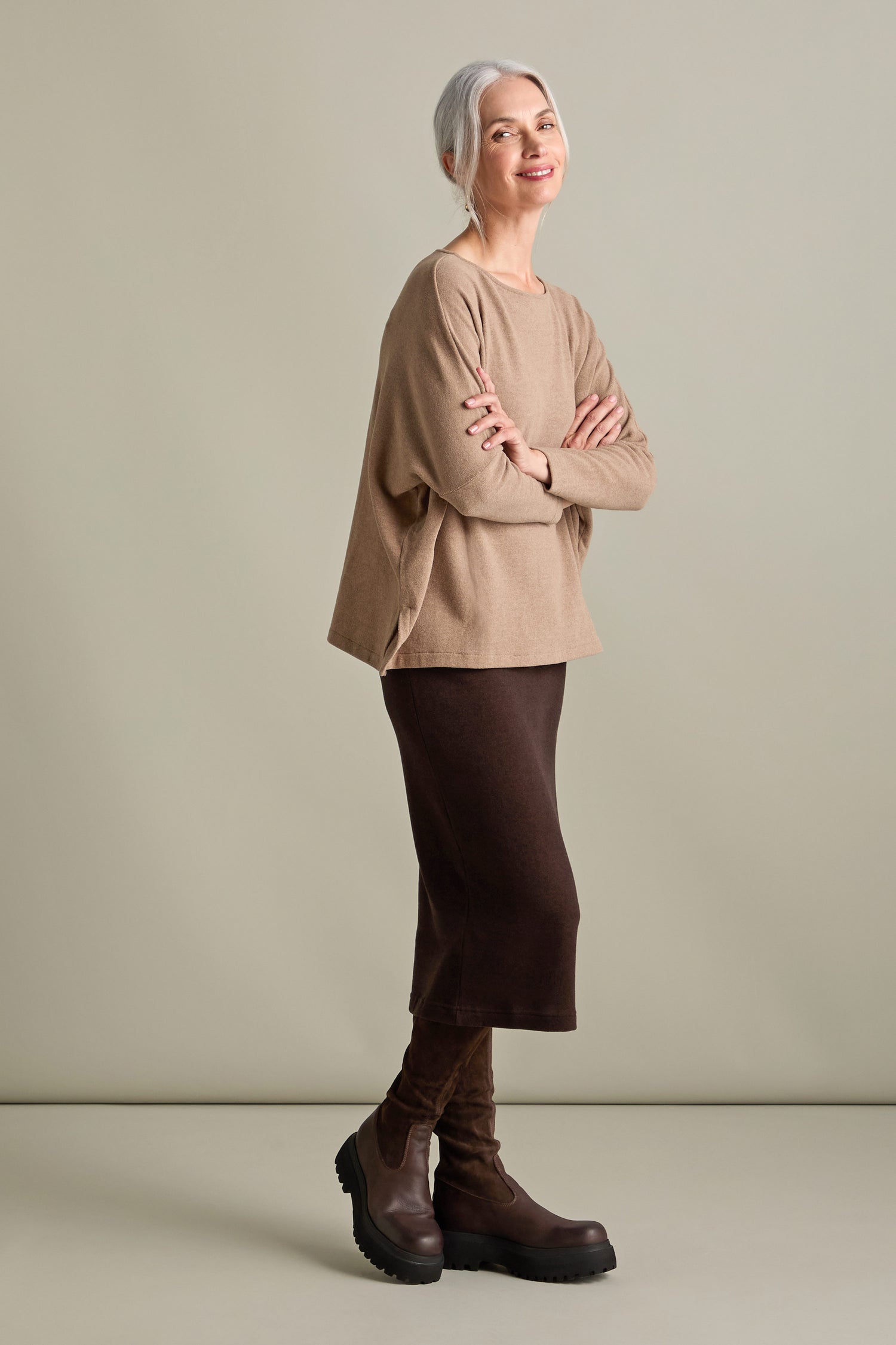 An older woman with gray hair stands with her arms crossed, wearing a beige sweater, dark brown skirt, and brown boots. She smiles with a relaxed posture against a plain background, donning a Martis Scoop Neck Soft Jersey Top made in Italy.