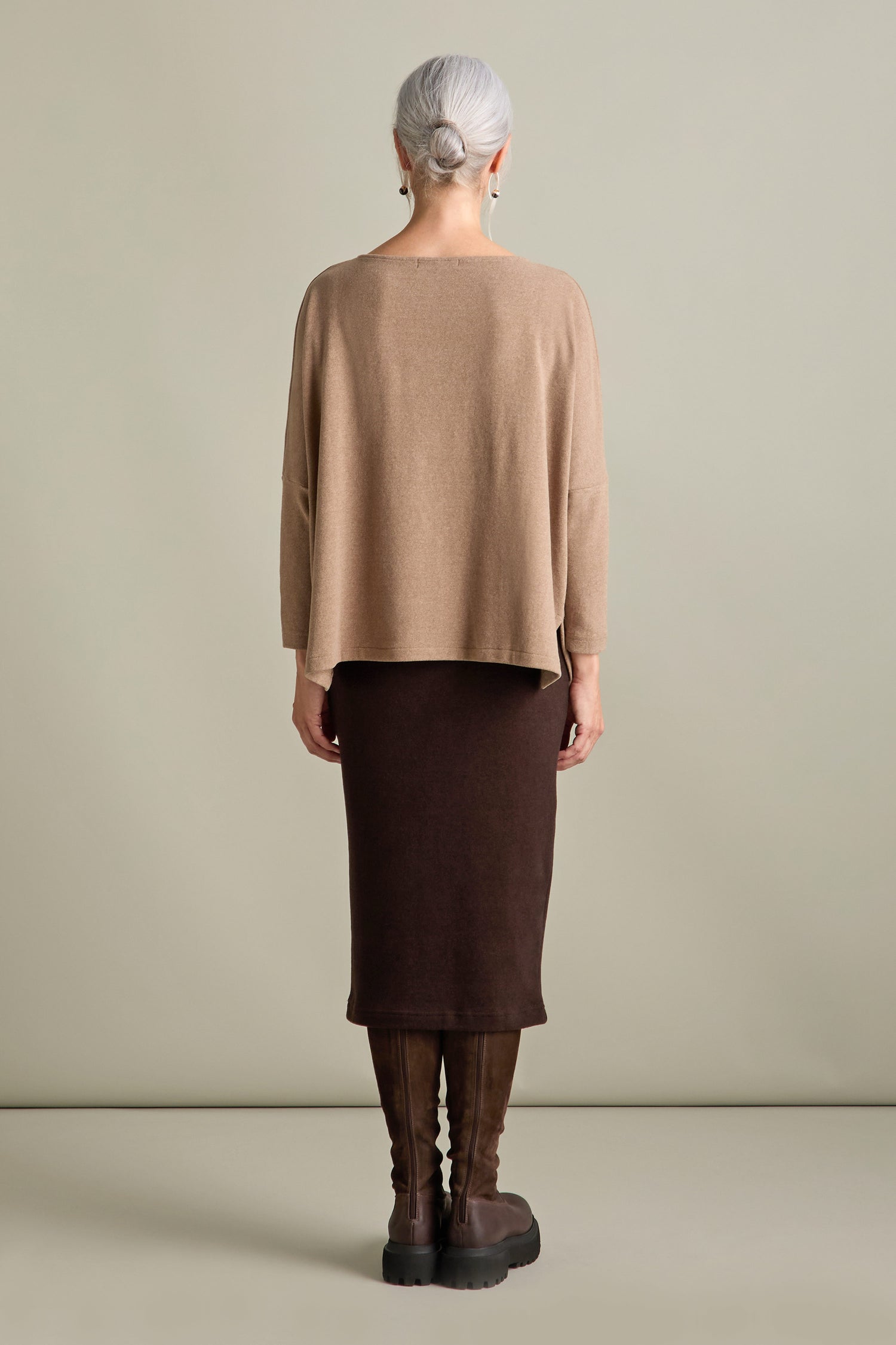 A person with gray hair, dressed in a Martis Scoop Neck Soft Jersey Top from Mama b, made in Italy, a brown skirt, and knee-high boots, is standing with their back to the camera against a plain background.