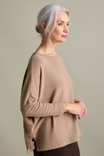 A woman with grey hair is standing sideways, wearing a beige long-sleeved Martis Scoop Neck Soft Jersey Top made in Italy, and looking forward.