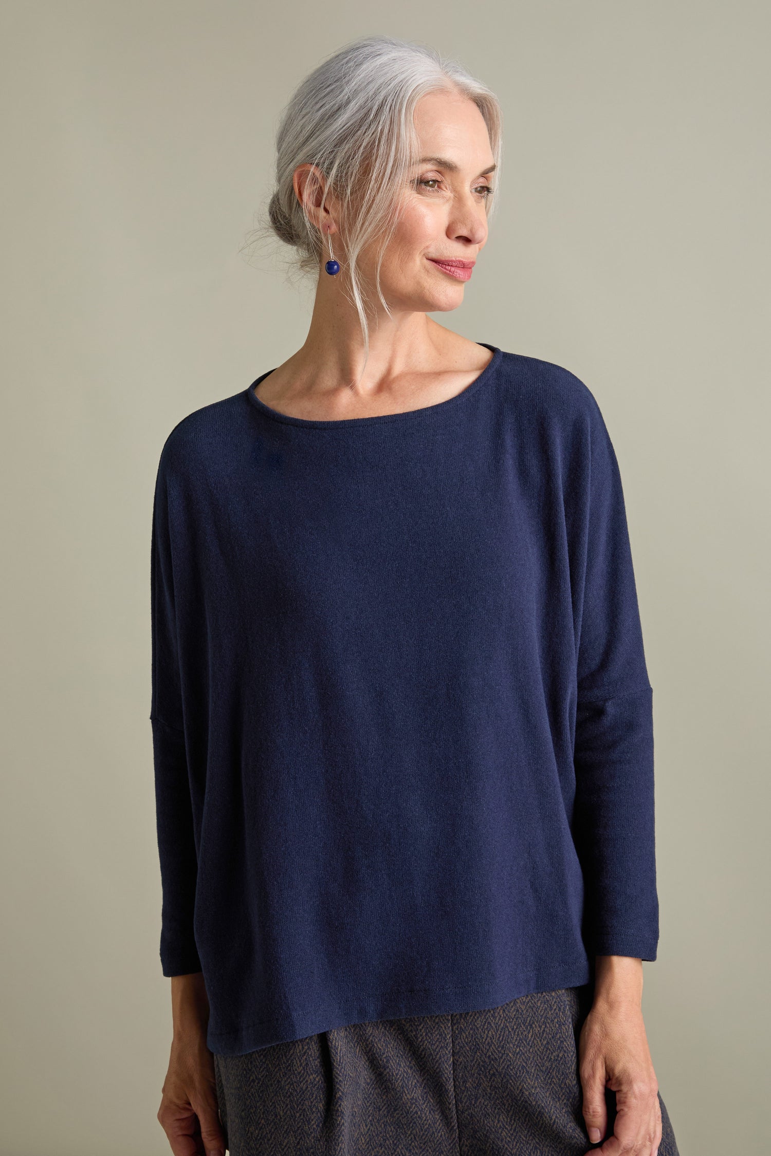 An older woman with gray hair, wearing a blue Martis Scoop Neck Soft Jersey Top made in Italy and dark pants, stands against a neutral background, facing slightly to the left.