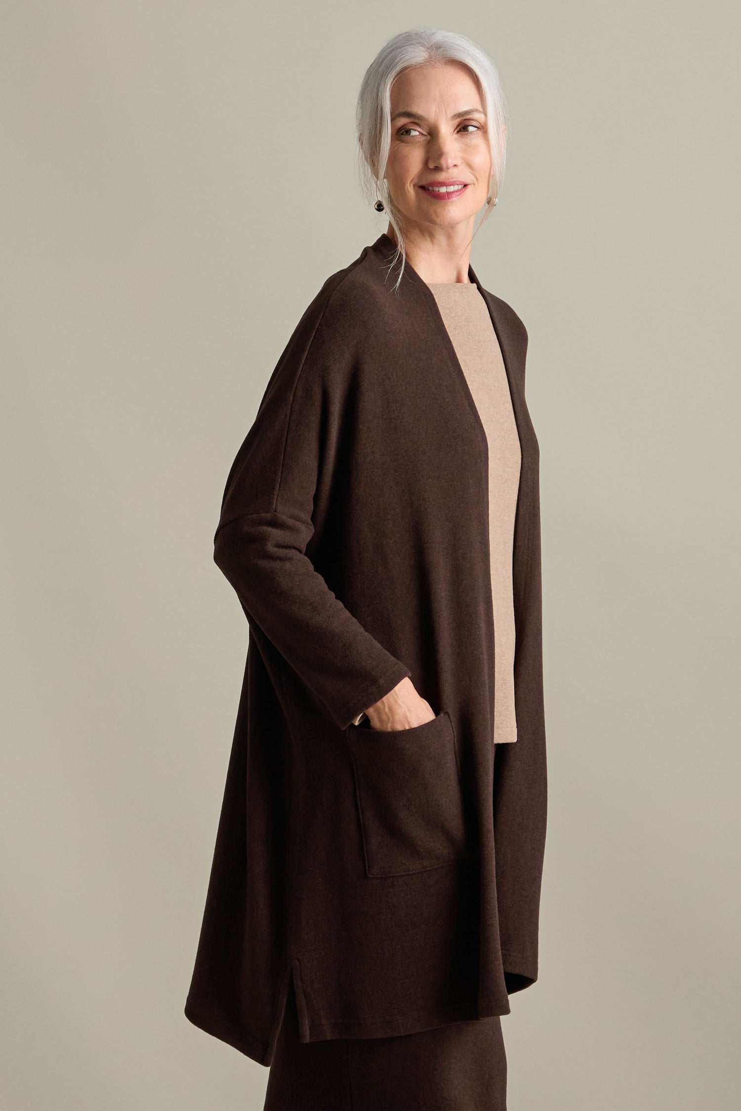 A woman with gray hair is standing, wearing a Souffle Soft Jersey Cardi in longline length brown over a beige top. She has her hands in the cardigan pockets and is looking off to the side while smiling.