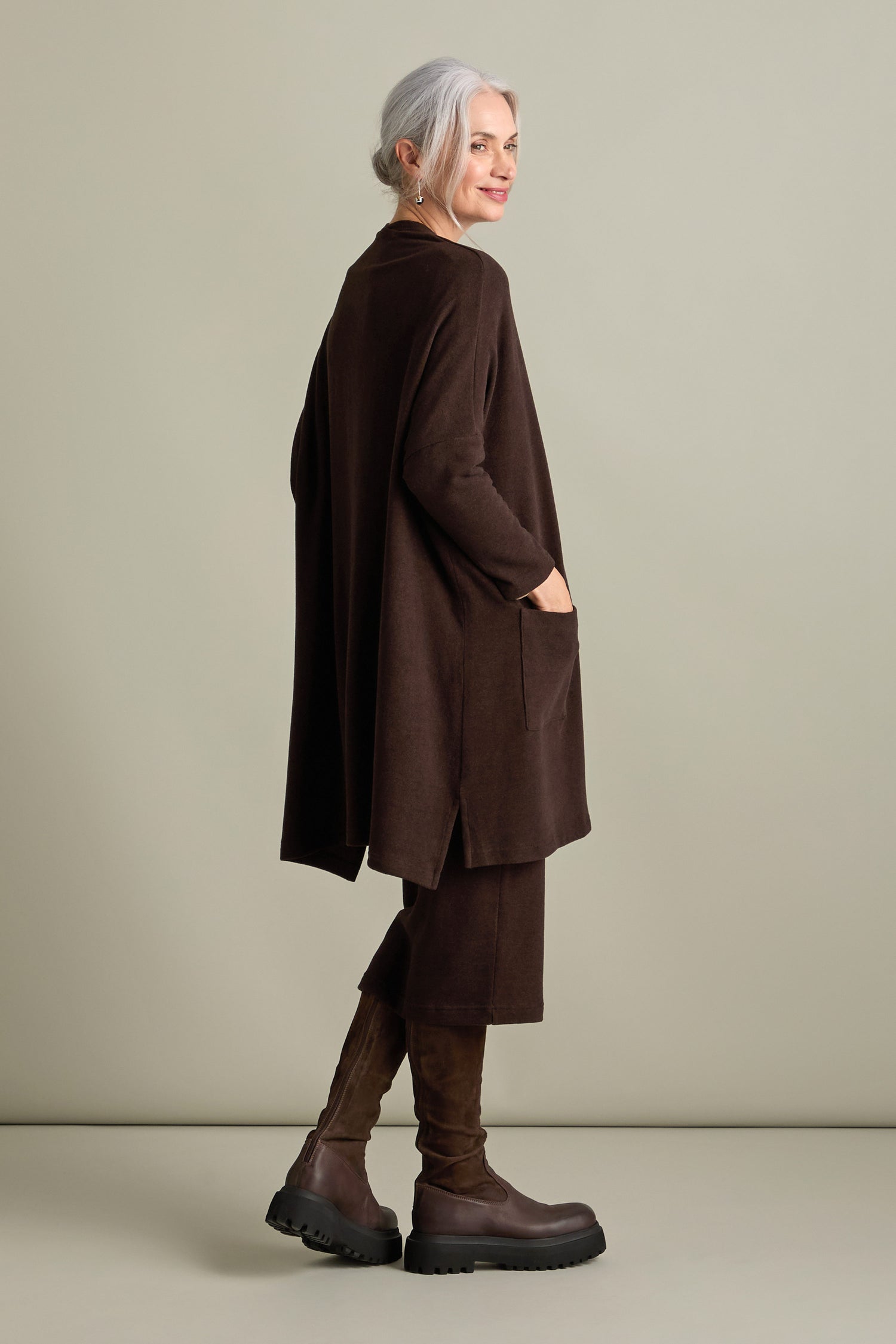 A woman with gray hair stands and turns slightly to her left, dressed in a longline brown Souffle Soft Jersey Cardi by Mama b, matching pants, and knee-high boots, against a plain background.