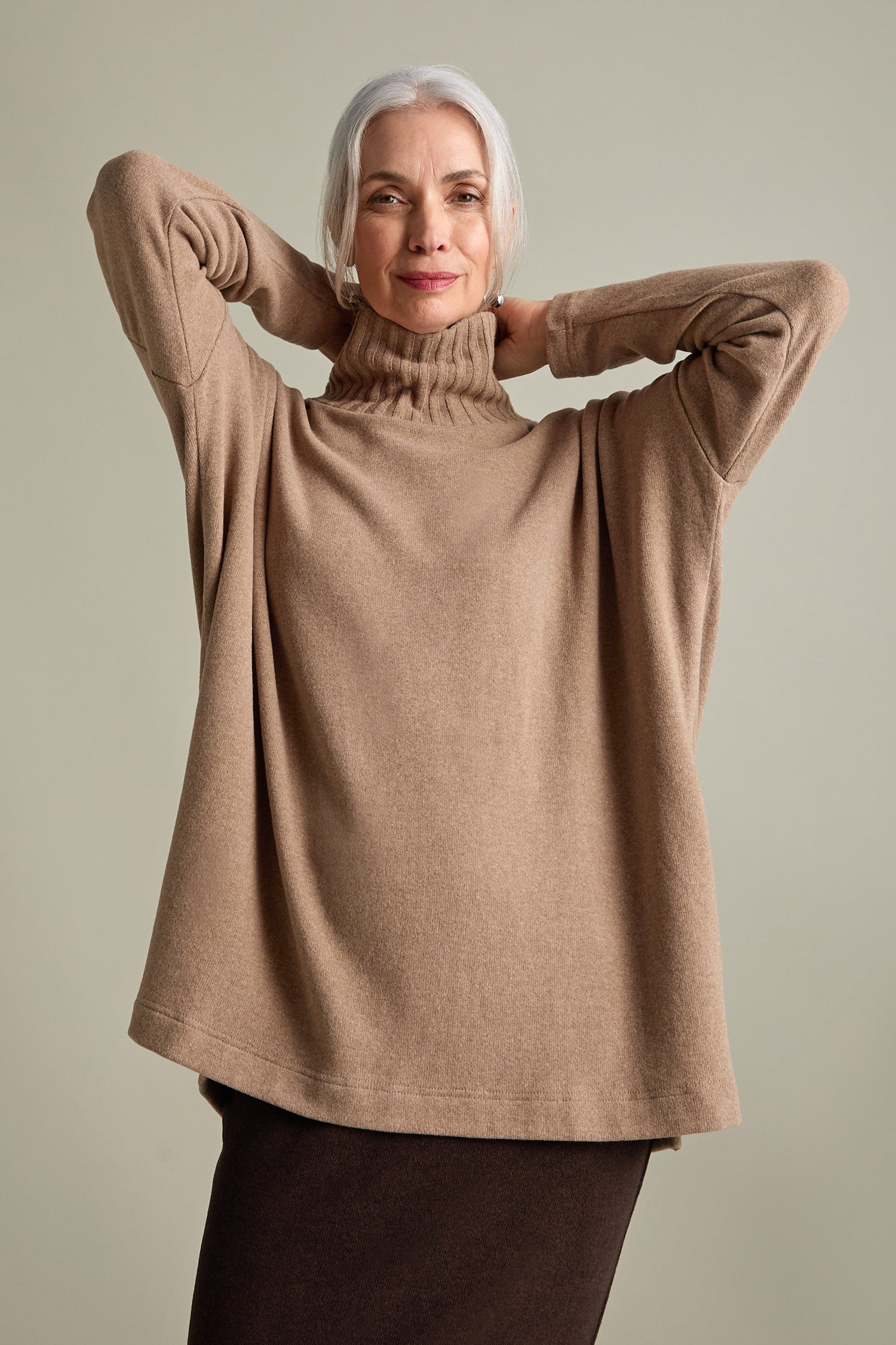 A woman with gray hair is wearing a Zeppola Rib Neck Soft Jersey Tunic and a dark skirt, standing with her hands behind her head against a plain background.