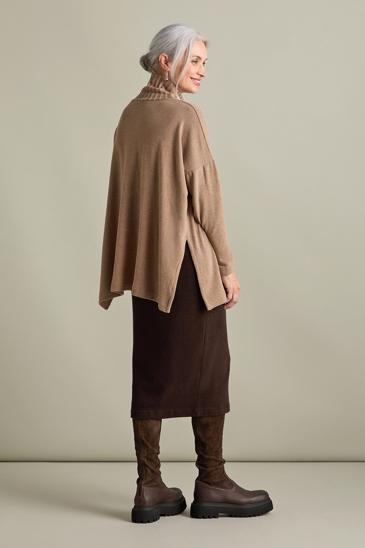 A person with gray hair is standing and smiling while wearing a cozy Zeppola Rib Neck Soft Jersey Tunic, a brown skirt, and dark brown knee-high boots. The background is a plain light beige color.