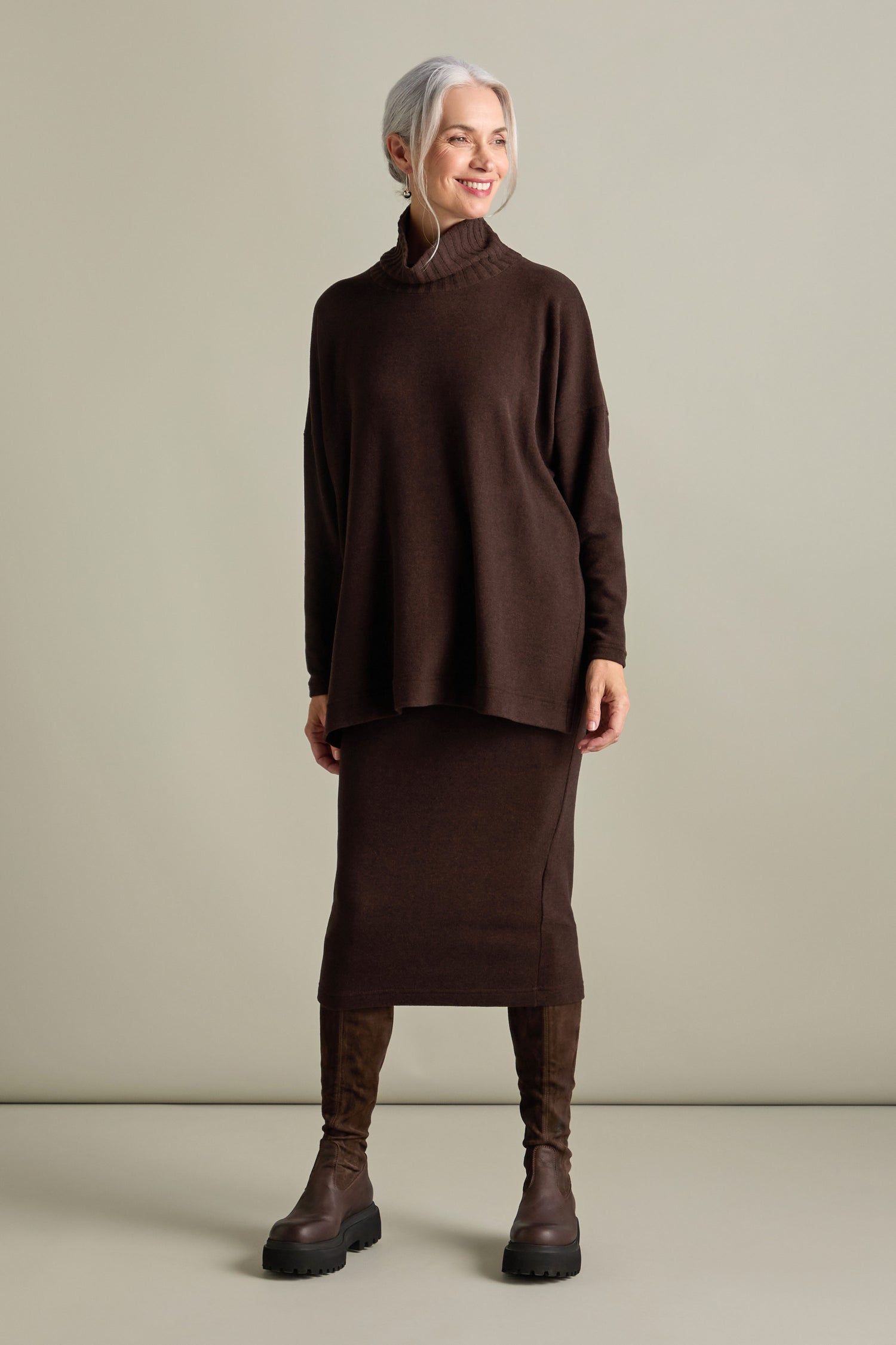 A person with gray hair in a ponytail wears a loose, dark brown Zeppola Rib Neck Soft Jersey Tunic with a high neckline, paired with a matching long skirt and brown boots. They are standing in a neutral-colored room and smiling.