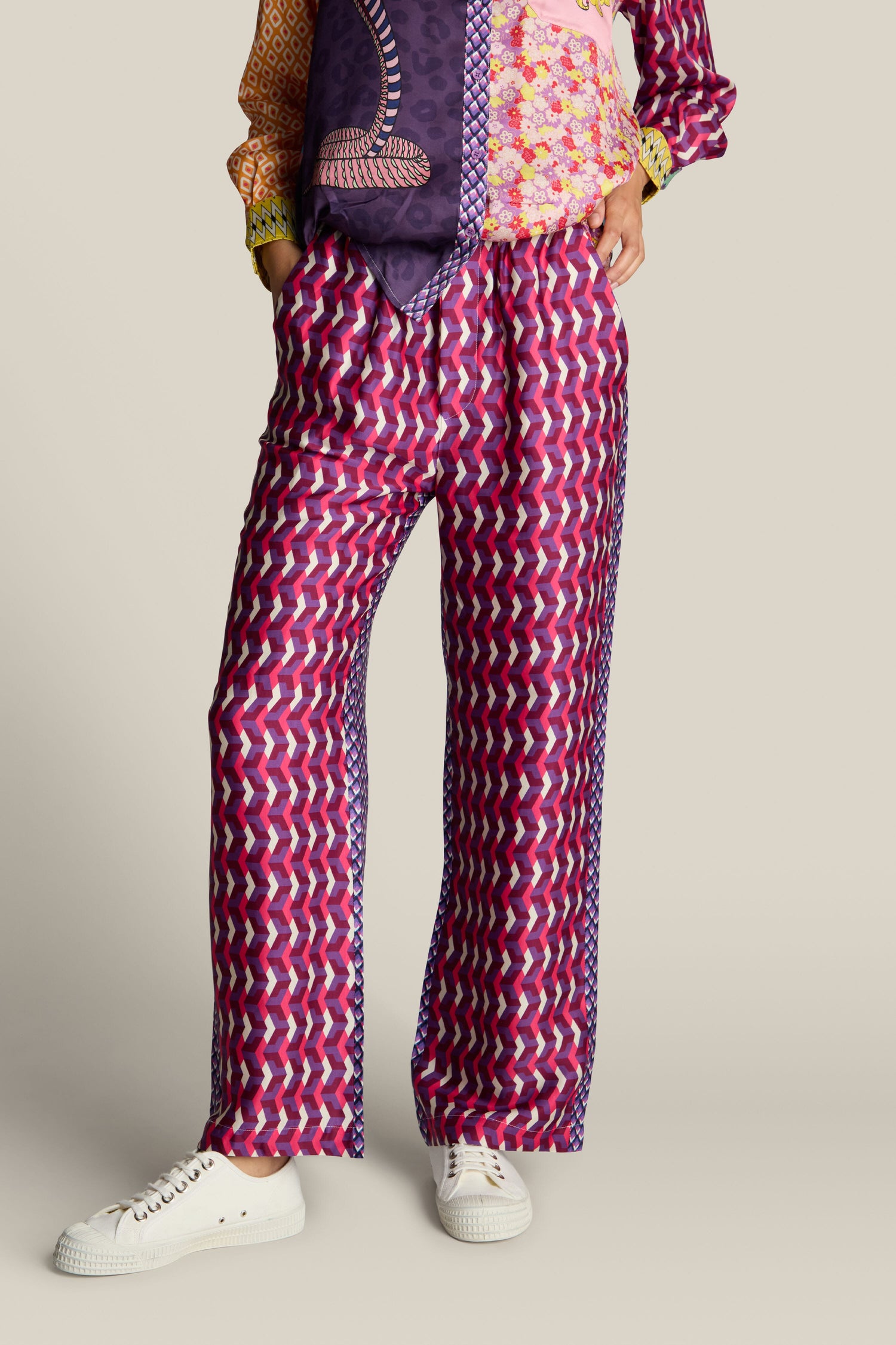 A person wearing the Twisted Love Bailey Trouser in a vibrant mix of purple, pink, and white, paired with white sneakers and a colorful long-sleeve shirt.