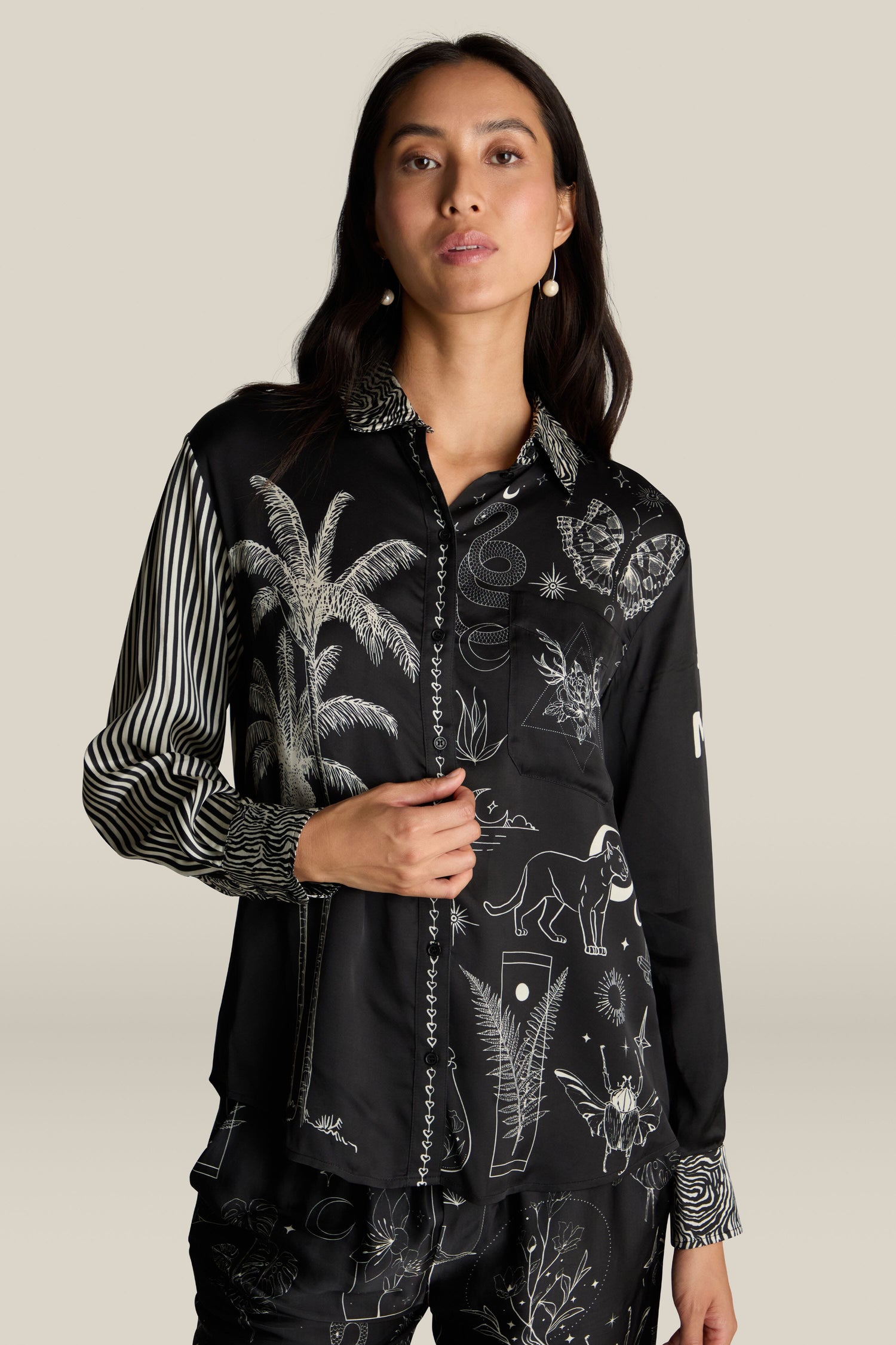 A woman with long dark hair faces forward against a plain background, wearing the Black Magic Isabel Shirt, which features a black button-up design adorned with intricate white illustrations and striped sleeves