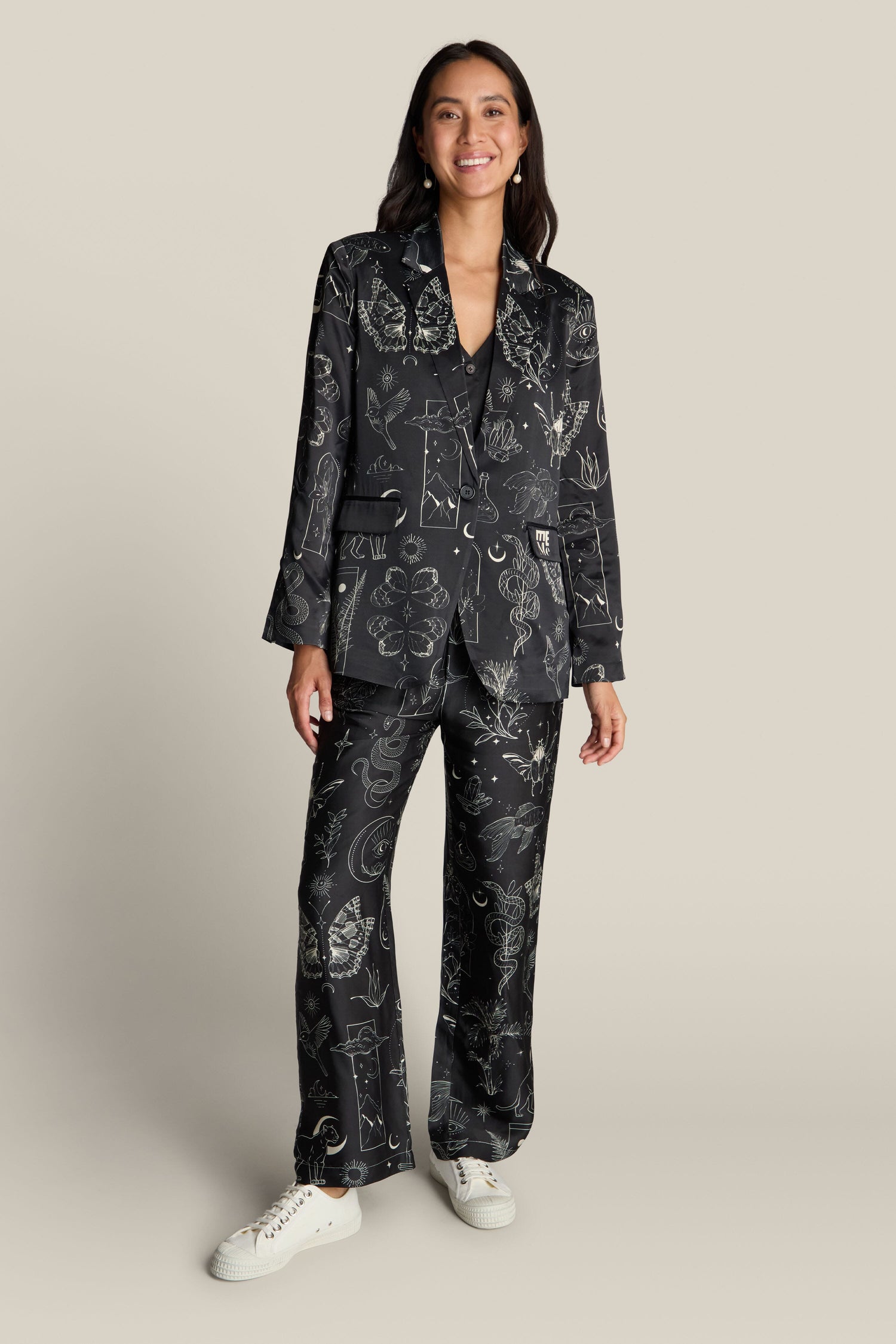 A woman is standing and wearing the Molly Printed Jacket crafted from luxurious viscose and rayon, featuring a mystical print. She has long hair and is smiling, effortlessly paired with white sneakers against a plain beige background.