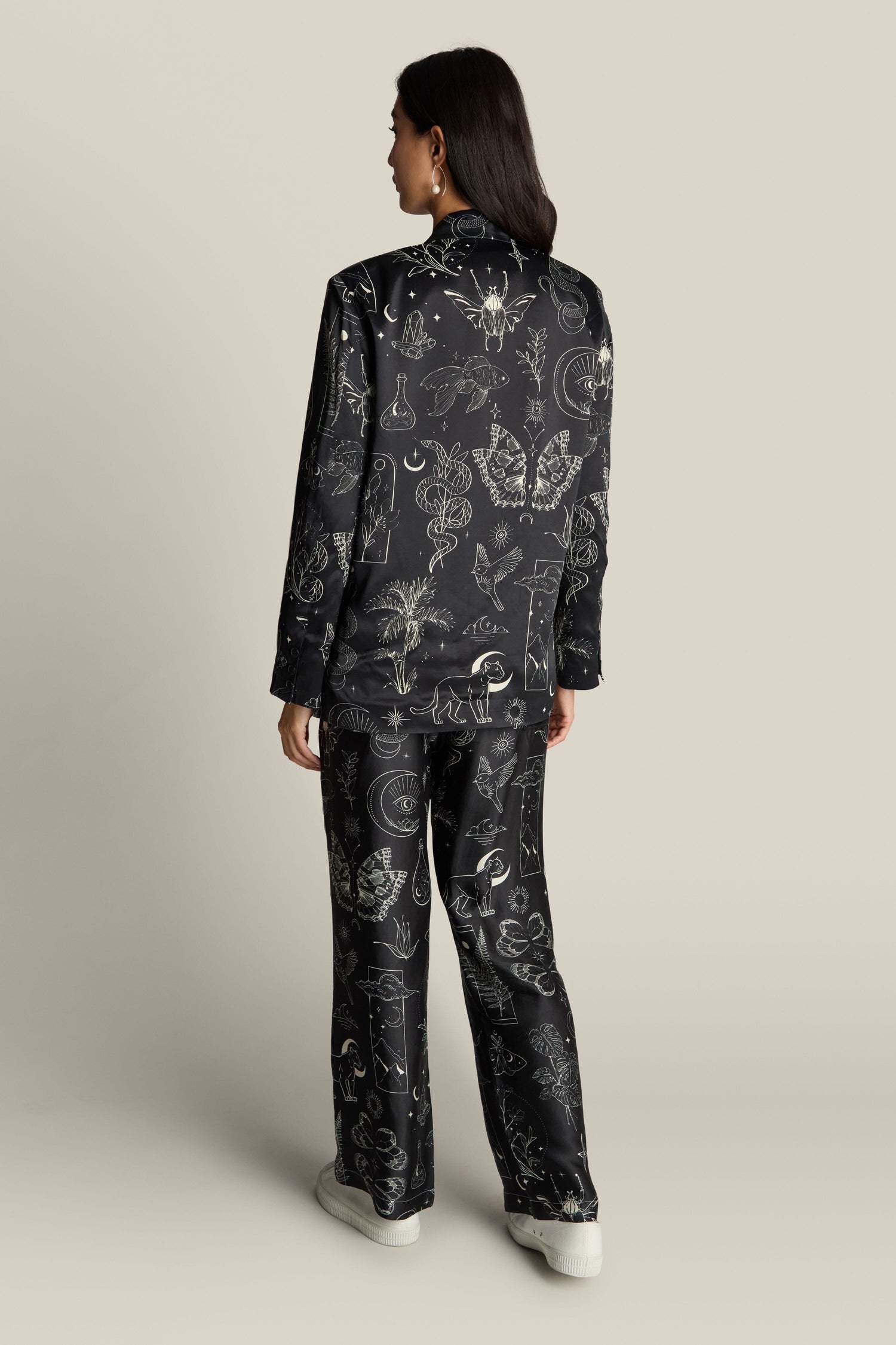 A person stands with their back turned, clad in the Molly Printed Jacket, a black patterned pantsuit adorned with various white designs and hints of mystical prints, set against a plain beige background.