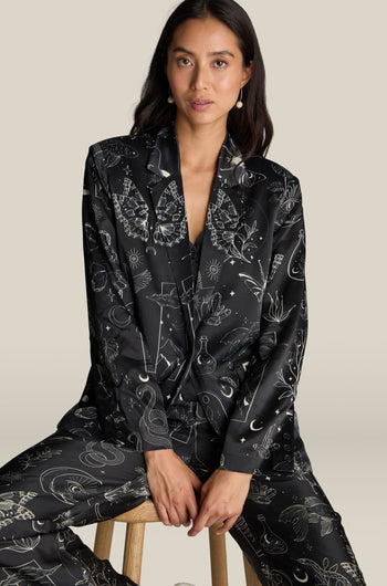 A woman wearing a black patterned suit, including the luxurious Molly Printed Jacket adorned with a mystical print, sits on a stool against a plain background.