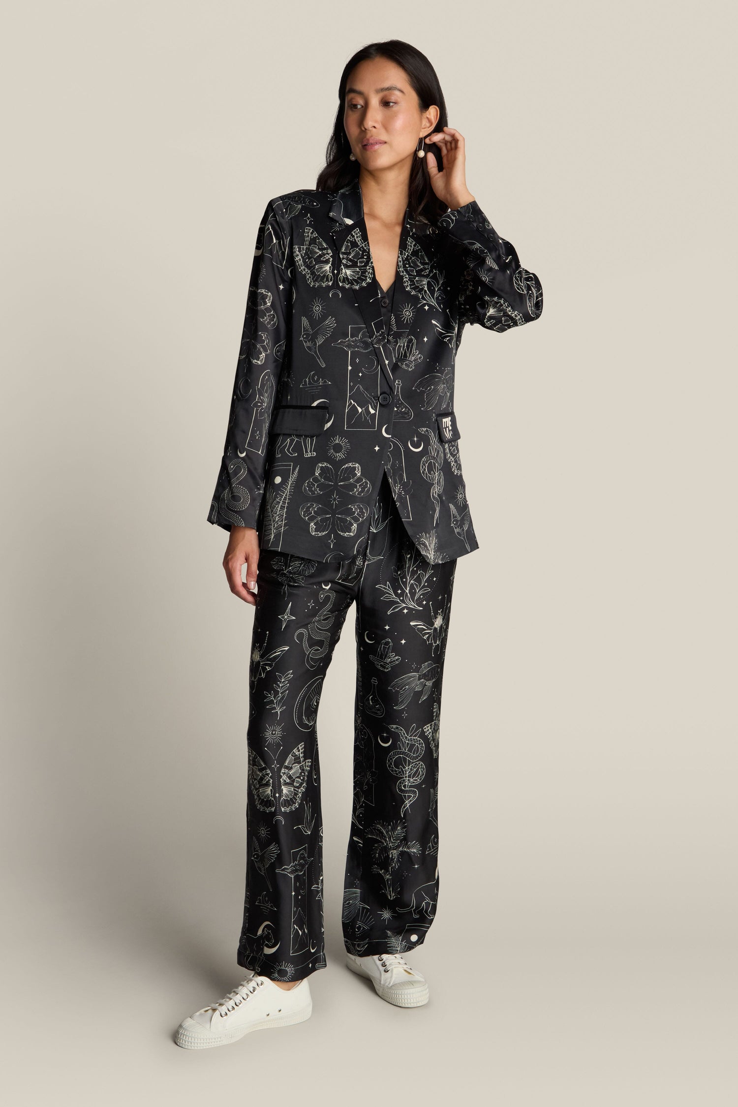 A person stands against a plain background wearing the luxurious Molly Printed Jacket, crafted from viscose and rayon fabric, which features an enchanting mystical print. They pair the jacket with matching pants, white sneakers, and don a relaxed hairstyle.