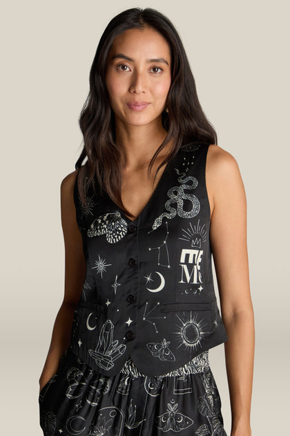 A woman is wearing a Cameron Vest embellished with white celestial and mystical digital prints, featuring a snake, crescent moons, and geometric shapes. She has long dark hair and stands against a plain background.