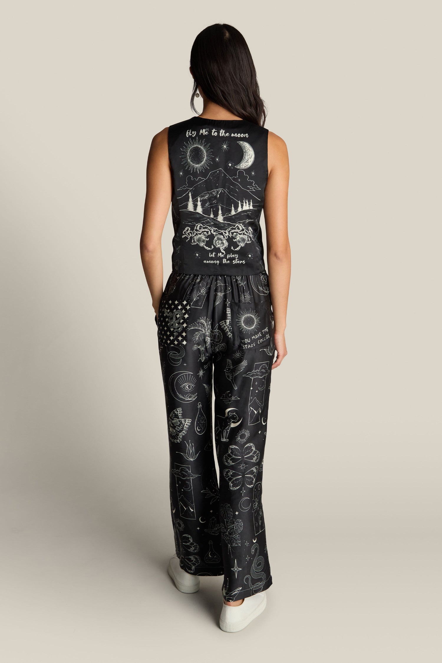 A person with long dark hair is standing with their back to the camera, wearing the Cameron Vest and black pants featuring various mystical digital print illustrations and text.