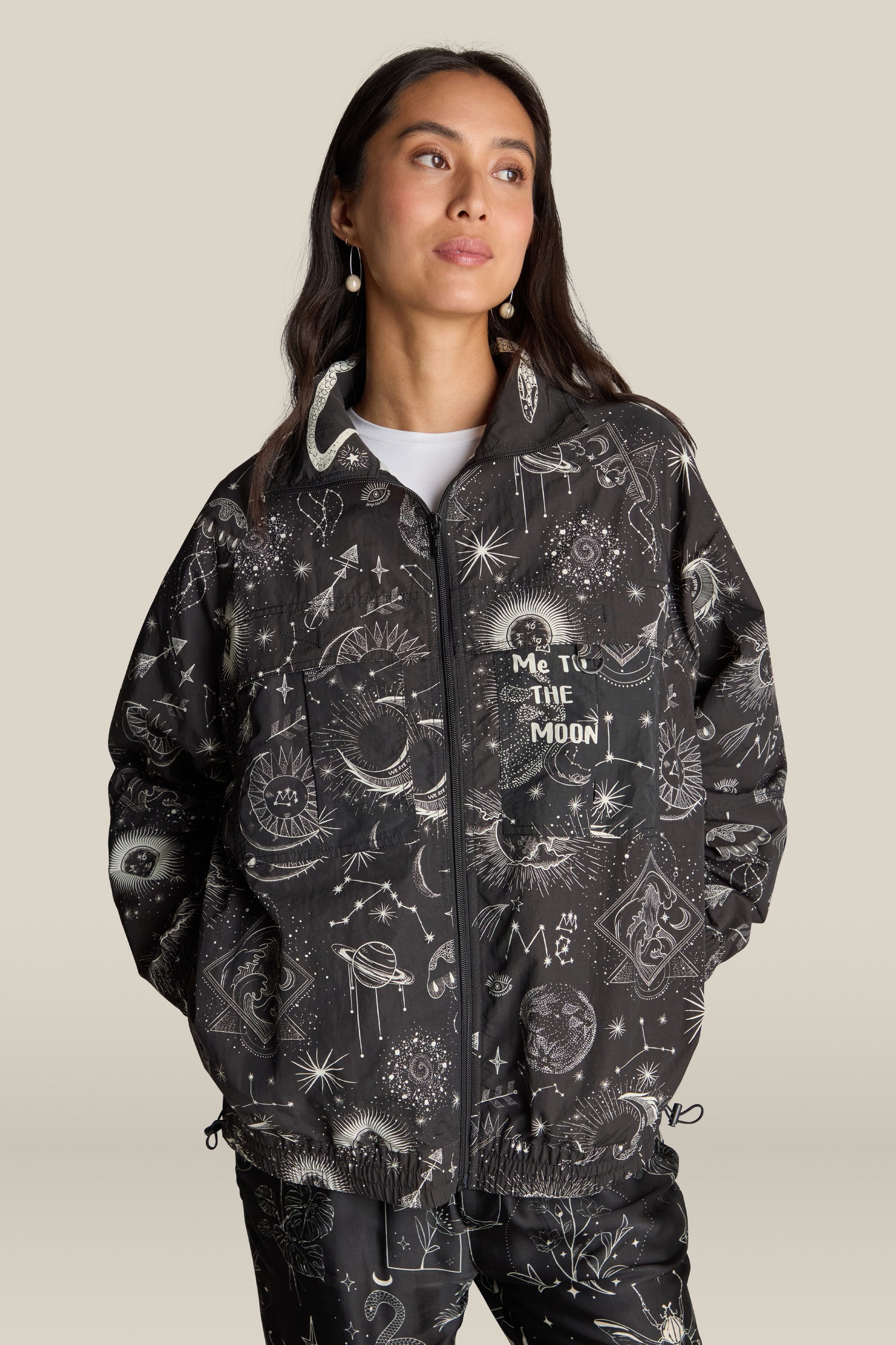 A woman stands with hands in pockets, donning a celestial-themed Jolene Track Jacket and matching pants. The jacket, emblazoned with "Me to the Moon," features a stunning constellation print. She gazes to her right, set against a neutral background, epitomizing the ME369 style.