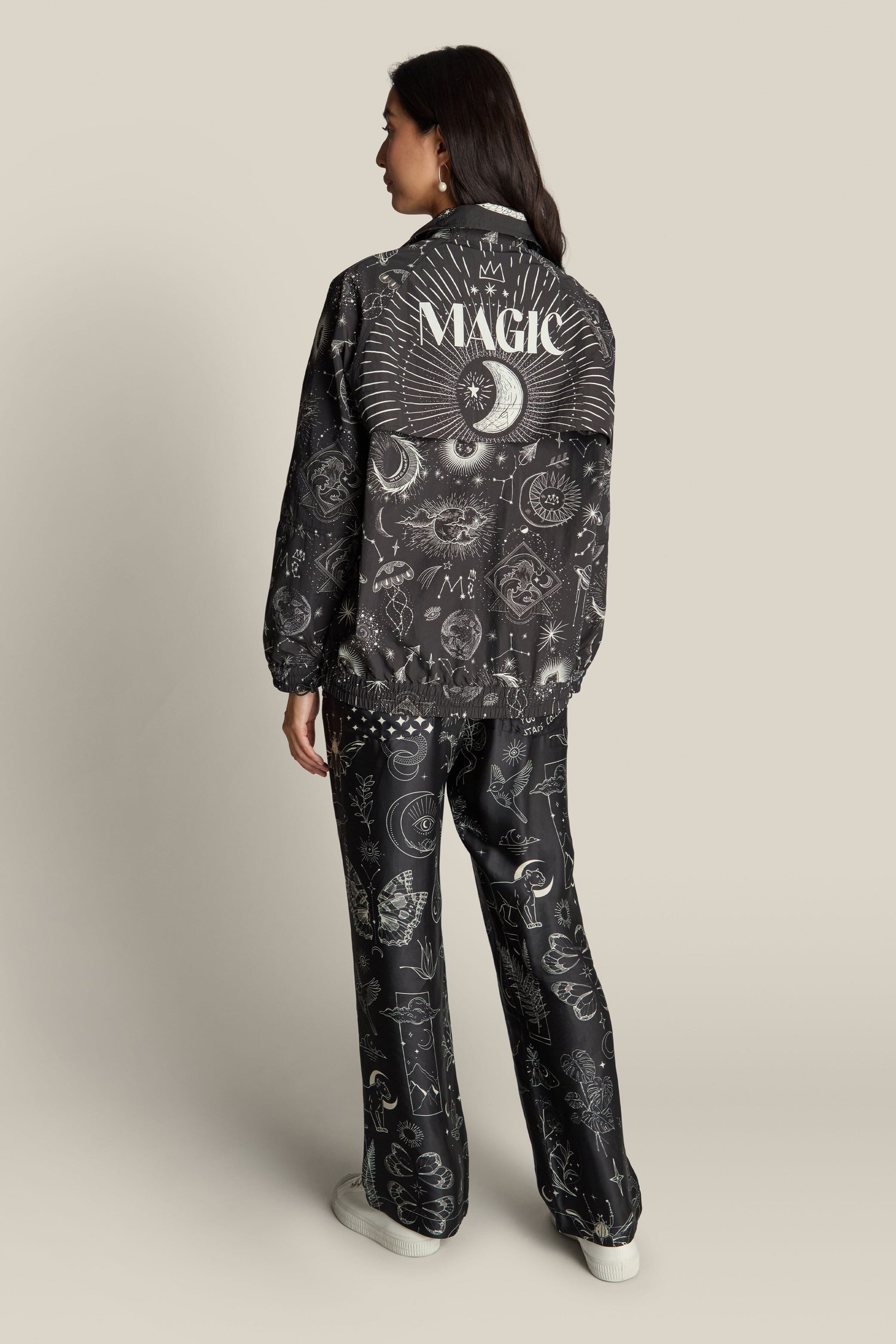 A person facing away, wearing a black Jolene Track Jacket adorned with mystical symbols and "MAGIC" written on the back. They are paired with matching black pants featuring constellation print designs.