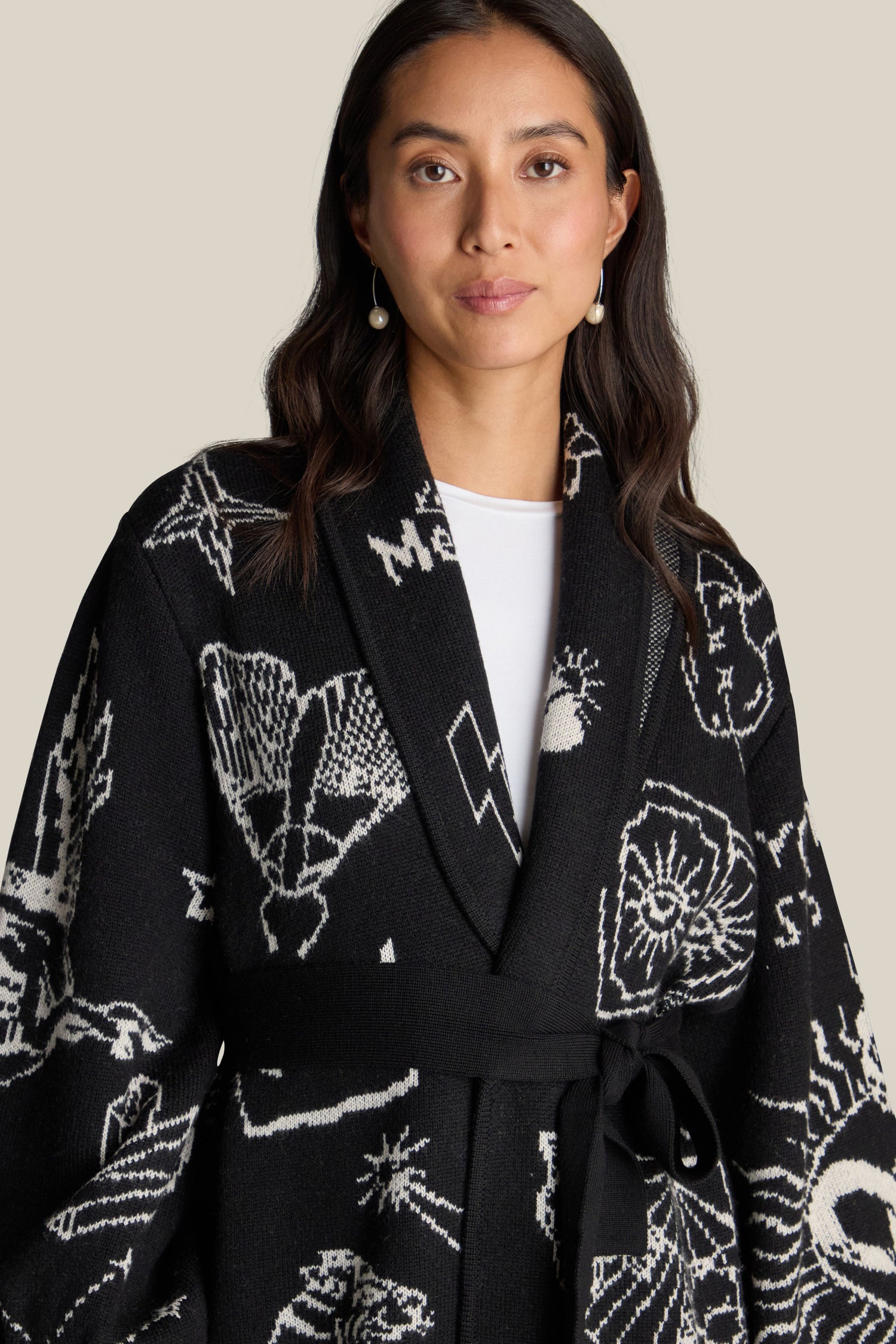 A woman with long dark hair wears the Barbara Long Cardigan, a black and white patterned wrap coat with a belt. The 100% wool piece features a mystical design that complements her pearl earrings as she stands against a plain beige background.