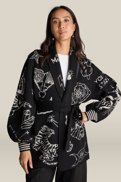 A person stands confidently wearing the Barbara Long Cardigan in black, featuring a belt and crafted from 100% wool, adorned with various white illustrations and symbols that lend a mystical design.