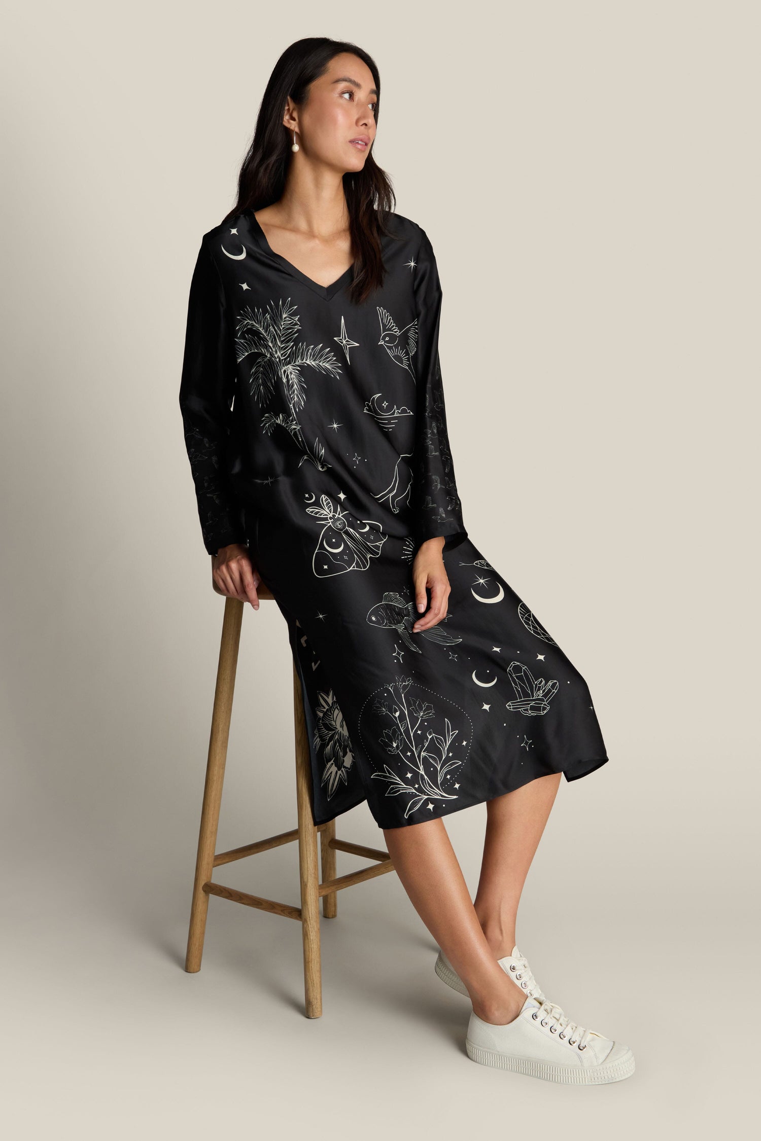 A woman sits on a wooden stool wearing the Violet Dress, featuring long sleeves adorned with white celestial and botanical designs, paired with white sneakers. The deep V-neck adds an elegant touch to her ensemble.